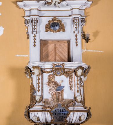 Zdjęcie nr 11: Ambona zawieszona jest na ścianie, z wejściem w zaplecku. Kosz ambony, na rzucie owalu, artykułują cztery pilastry osadzone na obiegającym jego podstawę cokole, wspierające wyłamujący się ponad nimi gzyms wieńczący. Podwieszenie kosza jest w formie czterech wolutowych kabłąków spiętych spłaszczonym pierścieniem. Zaplecek jest w formie prostokątnej płyciny ujętej parą pilastrów wspierających wyłamujące się ponad nimi belkowanie z wydatnym gzymsem. Zwieńczenie ma formę półkolistego przyczółka. Pola kosza ambony wypełniają płaskorzeźbione przedstawienia trzech świętych Janów. W środkowym znajduje się św. Jan Kanty siedzący przy pulpicie z otwartą księgą, poniżej znajduje się

kartusz herbowy, o krawędziach zdobionych ornamentyką akantową, z napisem: „Et lex / clementia / in linqua / eius”, natomiast w medalionie powyżej jest zamknięta księga z siedmioma pieczęciami. Po lewej znajduje się św. Jan Ewangelista, poniżej na kartuszu napis: „Quia verum est testimo/nium eius”, w medalionie zaś klatka. Po prawej jest przedstawienie św. Jana Chrzciciela z kartuszem z napisem: „Et enim / manus Domini / errat /cum / suo” oraz medalionem ze znakiem ręki trzymającej trąbę. Pilastry zdobią motywy kampanulli oraz uskrzydlonych główek anielskich, z których niektóre są na tle konch. Tuż pod gzymsem wieńczącym podwieszona jest girlanda kwiatowa. Belkowanie zaplecka ambony wsparte jest dodatkowo na wolutowych wspornikach, trzony pilastrów zdobią kampanulle. Po środku płyciny zaplecka, nad wejściem znajduje się ujęty akantem kartusz herbowy z napisem: „Docebit vos omnia”. Zwieńczenie ambony zdobią figurki dwóch par aniołków, z czego jedna usytuowana jest na krawędziach przyczółka, druga wypełnia pole tympanonu. Ukazane naprzeciw siebie w pozycjach półleżących, o przeplatających się nogach obuczącz podtrzymują otwartą księgę z napisem: „EVANGELIUM IESU CHRISTI”. Szczyt zwieńczenia jest w formie promienistej glorii z gołębicą Ducha Świętego po środku. Struktura ambony jest pomalowana na biało, podobnie, jak odsłonięte partie ciał aniołków. Detal architektoniczny, ornamnetyka, szaty i skrzydła są złocone i srebrzone.