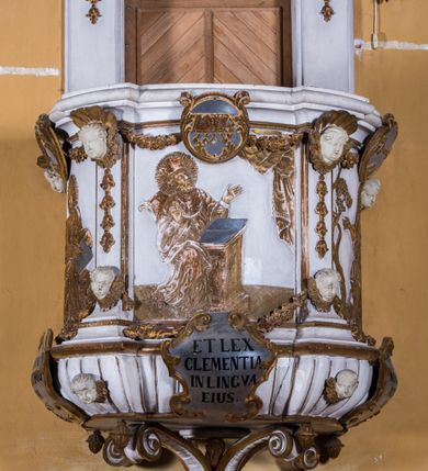 Zdjęcie nr 12: Ambona zawieszona jest na ścianie, z wejściem w zaplecku. Kosz ambony, na rzucie owalu, artykułują cztery pilastry osadzone na obiegającym jego podstawę cokole, wspierające wyłamujący się ponad nimi gzyms wieńczący. Podwieszenie kosza jest w formie czterech wolutowych kabłąków spiętych spłaszczonym pierścieniem. Zaplecek jest w formie prostokątnej płyciny ujętej parą pilastrów wspierających wyłamujące się ponad nimi belkowanie z wydatnym gzymsem. Zwieńczenie ma formę półkolistego przyczółka. Pola kosza ambony wypełniają płaskorzeźbione przedstawienia trzech świętych Janów. W środkowym znajduje się św. Jan Kanty siedzący przy pulpicie z otwartą księgą, poniżej znajduje się

kartusz herbowy, o krawędziach zdobionych ornamentyką akantową, z napisem: „Et lex / clementia / in linqua / eius”, natomiast w medalionie powyżej jest zamknięta księga z siedmioma pieczęciami. Po lewej znajduje się św. Jan Ewangelista, poniżej na kartuszu napis: „Quia verum est testimo/nium eius”, w medalionie zaś klatka. Po prawej jest przedstawienie św. Jana Chrzciciela z kartuszem z napisem: „Et enim / manus Domini / errat /cum / suo” oraz medalionem ze znakiem ręki trzymającej trąbę. Pilastry zdobią motywy kampanulli oraz uskrzydlonych główek anielskich, z których niektóre są na tle konch. Tuż pod gzymsem wieńczącym podwieszona jest girlanda kwiatowa. Belkowanie zaplecka ambony wsparte jest dodatkowo na wolutowych wspornikach, trzony pilastrów zdobią kampanulle. Po środku płyciny zaplecka, nad wejściem znajduje się ujęty akantem kartusz herbowy z napisem: „Docebit vos omnia”. Zwieńczenie ambony zdobią figurki dwóch par aniołków, z czego jedna usytuowana jest na krawędziach przyczółka, druga wypełnia pole tympanonu. Ukazane naprzeciw siebie w pozycjach półleżących, o przeplatających się nogach obuczącz podtrzymują otwartą księgę z napisem: „EVANGELIUM IESU CHRISTI”. Szczyt zwieńczenia jest w formie promienistej glorii z gołębicą Ducha Świętego po środku. Struktura ambony jest pomalowana na biało, podobnie, jak odsłonięte partie ciał aniołków. Detal architektoniczny, ornamnetyka, szaty i skrzydła są złocone i srebrzone.