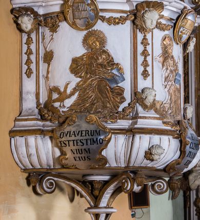 Zdjęcie nr 13: Ambona zawieszona jest na ścianie, z wejściem w zaplecku. Kosz ambony, na rzucie owalu, artykułują cztery pilastry osadzone na obiegającym jego podstawę cokole, wspierające wyłamujący się ponad nimi gzyms wieńczący. Podwieszenie kosza jest w formie czterech wolutowych kabłąków spiętych spłaszczonym pierścieniem. Zaplecek jest w formie prostokątnej płyciny ujętej parą pilastrów wspierających wyłamujące się ponad nimi belkowanie z wydatnym gzymsem. Zwieńczenie ma formę półkolistego przyczółka. Pola kosza ambony wypełniają płaskorzeźbione przedstawienia trzech świętych Janów. W środkowym znajduje się św. Jan Kanty siedzący przy pulpicie z otwartą księgą, poniżej znajduje się

kartusz herbowy, o krawędziach zdobionych ornamentyką akantową, z napisem: „Et lex / clementia / in linqua / eius”, natomiast w medalionie powyżej jest zamknięta księga z siedmioma pieczęciami. Po lewej znajduje się św. Jan Ewangelista, poniżej na kartuszu napis: „Quia verum est testimo/nium eius”, w medalionie zaś klatka. Po prawej jest przedstawienie św. Jana Chrzciciela z kartuszem z napisem: „Et enim / manus Domini / errat /cum / suo” oraz medalionem ze znakiem ręki trzymającej trąbę. Pilastry zdobią motywy kampanulli oraz uskrzydlonych główek anielskich, z których niektóre są na tle konch. Tuż pod gzymsem wieńczącym podwieszona jest girlanda kwiatowa. Belkowanie zaplecka ambony wsparte jest dodatkowo na wolutowych wspornikach, trzony pilastrów zdobią kampanulle. Po środku płyciny zaplecka, nad wejściem znajduje się ujęty akantem kartusz herbowy z napisem: „Docebit vos omnia”. Zwieńczenie ambony zdobią figurki dwóch par aniołków, z czego jedna usytuowana jest na krawędziach przyczółka, druga wypełnia pole tympanonu. Ukazane naprzeciw siebie w pozycjach półleżących, o przeplatających się nogach obuczącz podtrzymują otwartą księgę z napisem: „EVANGELIUM IESU CHRISTI”. Szczyt zwieńczenia jest w formie promienistej glorii z gołębicą Ducha Świętego po środku. Struktura ambony jest pomalowana na biało, podobnie, jak odsłonięte partie ciał aniołków. Detal architektoniczny, ornamnetyka, szaty i skrzydła są złocone i srebrzone.