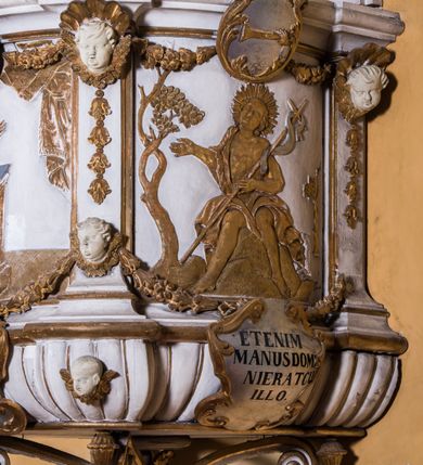 Zdjęcie nr 3: Ambona zawieszona jest na ścianie, z wejściem w zaplecku. Kosz ambony, na rzucie owalu, artykułują cztery pilastry osadzone na obiegającym jego podstawę cokole, wspierające wyłamujący się ponad nimi gzyms wieńczący. Podwieszenie kosza jest w formie czterech wolutowych kabłąków spiętych spłaszczonym pierścieniem. Zaplecek jest w formie prostokątnej płyciny ujętej parą pilastrów wspierających wyłamujące się ponad nimi belkowanie z wydatnym gzymsem. Zwieńczenie ma formę półkolistego przyczółka. Pola kosza ambony wypełniają płaskorzeźbione przedstawienia trzech świętych Janów. W środkowym znajduje się św. Jan Kanty siedzący przy pulpicie z otwartą księgą, poniżej znajduje się

kartusz herbowy, o krawędziach zdobionych ornamentyką akantową, z napisem: „Et lex / clementia / in linqua / eius”, natomiast w medalionie powyżej jest zamknięta księga z siedmioma pieczęciami. Po lewej znajduje się św. Jan Ewangelista, poniżej na kartuszu napis: „Quia verum est testimo/nium eius”, w medalionie zaś klatka. Po prawej jest przedstawienie św. Jana Chrzciciela z kartuszem z napisem: „Et enim / manus Domini / errat /cum / suo” oraz medalionem ze znakiem ręki trzymającej trąbę. Pilastry zdobią motywy kampanulli oraz uskrzydlonych główek anielskich, z których niektóre są na tle konch. Tuż pod gzymsem wieńczącym podwieszona jest girlanda kwiatowa. Belkowanie zaplecka ambony wsparte jest dodatkowo na wolutowych wspornikach, trzony pilastrów zdobią kampanulle. Po środku płyciny zaplecka, nad wejściem znajduje się ujęty akantem kartusz herbowy z napisem: „Docebit vos omnia”. Zwieńczenie ambony zdobią figurki dwóch par aniołków, z czego jedna usytuowana jest na krawędziach przyczółka, druga wypełnia pole tympanonu. Ukazane naprzeciw siebie w pozycjach półleżących, o przeplatających się nogach obuczącz podtrzymują otwartą księgę z napisem: „EVANGELIUM IESU CHRISTI”. Szczyt zwieńczenia jest w formie promienistej glorii z gołębicą Ducha Świętego po środku. Struktura ambony jest pomalowana na biało, podobnie, jak odsłonięte partie ciał aniołków. Detal architektoniczny, ornamnetyka, szaty i skrzydła są złocone i srebrzone.