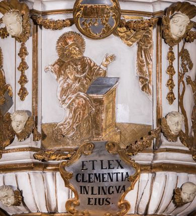 Zdjęcie nr 4: Ambona zawieszona jest na ścianie, z wejściem w zaplecku. Kosz ambony, na rzucie owalu, artykułują cztery pilastry osadzone na obiegającym jego podstawę cokole, wspierające wyłamujący się ponad nimi gzyms wieńczący. Podwieszenie kosza jest w formie czterech wolutowych kabłąków spiętych spłaszczonym pierścieniem. Zaplecek jest w formie prostokątnej płyciny ujętej parą pilastrów wspierających wyłamujące się ponad nimi belkowanie z wydatnym gzymsem. Zwieńczenie ma formę półkolistego przyczółka. Pola kosza ambony wypełniają płaskorzeźbione przedstawienia trzech świętych Janów. W środkowym znajduje się św. Jan Kanty siedzący przy pulpicie z otwartą księgą, poniżej znajduje się

kartusz herbowy, o krawędziach zdobionych ornamentyką akantową, z napisem: „Et lex / clementia / in linqua / eius”, natomiast w medalionie powyżej jest zamknięta księga z siedmioma pieczęciami. Po lewej znajduje się św. Jan Ewangelista, poniżej na kartuszu napis: „Quia verum est testimo/nium eius”, w medalionie zaś klatka. Po prawej jest przedstawienie św. Jana Chrzciciela z kartuszem z napisem: „Et enim / manus Domini / errat /cum / suo” oraz medalionem ze znakiem ręki trzymającej trąbę. Pilastry zdobią motywy kampanulli oraz uskrzydlonych główek anielskich, z których niektóre są na tle konch. Tuż pod gzymsem wieńczącym podwieszona jest girlanda kwiatowa. Belkowanie zaplecka ambony wsparte jest dodatkowo na wolutowych wspornikach, trzony pilastrów zdobią kampanulle. Po środku płyciny zaplecka, nad wejściem znajduje się ujęty akantem kartusz herbowy z napisem: „Docebit vos omnia”. Zwieńczenie ambony zdobią figurki dwóch par aniołków, z czego jedna usytuowana jest na krawędziach przyczółka, druga wypełnia pole tympanonu. Ukazane naprzeciw siebie w pozycjach półleżących, o przeplatających się nogach obuczącz podtrzymują otwartą księgę z napisem: „EVANGELIUM IESU CHRISTI”. Szczyt zwieńczenia jest w formie promienistej glorii z gołębicą Ducha Świętego po środku. Struktura ambony jest pomalowana na biało, podobnie, jak odsłonięte partie ciał aniołków. Detal architektoniczny, ornamnetyka, szaty i skrzydła są złocone i srebrzone.