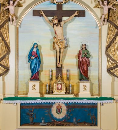 Zdjęcie nr 2: Przyścienny, architektoniczny ołtarz jednokondygnacyjny i jednoosiowy ze zwieńczeniem. Nastawa osadzona jest na cokole, w strukturze którego znajduje się prostopadłościenny ołtarz z drewnianą mensą i polichromowanym antepedium. Część środkowa retabulum, o kształcie zbliżonym do stojącego prostokąta zamkniętego trójkątnym szczytem, ujęta jest parą pilastrów osadzonych na cokołach. Zwieńczenie ołtarza przybiera formę ażurowej latarni na rzucie eliptycznym, ujętej dołem wyłamującym się gzymsem. Szczyt jest w kształcie zamkniętej, złoconej korony, wspartej na marmoryzowanych kolumienkach. Pod zwieńczenie ołtarza podwieszone jest złocone paludamentum podwiązane w górnych narożach części środkowej retabulum oraz podtrzymywane przez parę puttów i kaskadowo opadające wzdłuż pilastrów. Jego obrzeża zdobi frędzla oraz chwosty. Dodatkowo strukturę w górnej partii ołtarza zdobią: pośrodku motyw promienistej glorii z monogramem IHS z krzyżem i gorejącym sercem oraz motywy kogucich grzebieni po bokach. Pilastry zwieńczone są figurkami aniołów trzymających lichtarze, antepedium zdobi polichromowany motyw gorejącego serca Jezusa ujęty promienistą glorią oraz kłosami zbóż i gałązkami winnej latorośli, wraz z symbolami narzędzi męki Pańskiej (od lewej: kolumna biczowania i bicze, kogut, dzban z misą, młotek, obcęgi, gwoździe, lampa, rękawica, maczuga, miecz, kości do gry, trzciny, gąbka i włócznia). Pole środkowe, ujęte profilowaną, złoconą ramą, jest w kształcie leżącego prostokąta zamkniętego łukiem półkolistym. Zawiera rzeźbiarskie przedstawienie Ukrzyżowania z krucyfiksem ujętym po bokach parą figur asystencyjnych – Matki Boskiej i św. Jana Ewangelisty, ustawionych na cokołach. 
Struktura ołtarza jest utrzymana w odcieniu kremowym, partie detalu architektonicznego oraz ornamentyka są złocone i marmoryzowane w odcieniach brązu. 
 