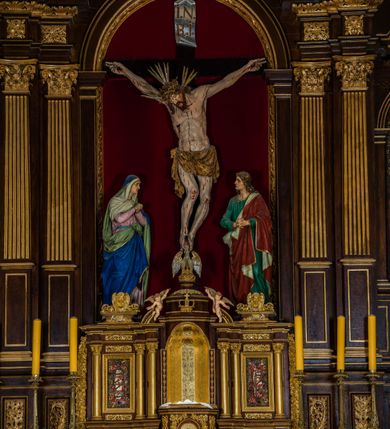 Zdjęcie nr 24: Ołtarz drewniany, architektoniczny, trójosiowy, jednokondygnacyjny ze zwieńczeniem usytuowany w połowie prezbiterium, na całej jego szerokości. Ołtarz prostopadłościenny z drewnianą mensą. Tabernakulum jest trójosiowe, osadzone na podwójnym cokole, zryzalitowane w części środkowej, podzielone kompozytowymi kolumnami, podtrzymującymi wyłamujące się nad nimi belkowanie z przerwanym przyczółkiem pośrodku. W części środkowej ruchomy tron wystawienia, wieloboczny, z rytymi literami IHS na tle glorii promienistej w jednej części i gałązkami z kwiatami w drugiej. Retabulum osadzone na wysokim cokole, z bramkami po bokach zamkniętymi łukami dwuramiennymi. Na dwiczkach malowane wizerunki św. Piotra po prawej i św. Pawła po lewej stronie. Część środkową retabulum ujmują dwie pary kompozytowych pilastrów o kanelowanych trzonech, z których środkowe cofnięte, podtrzymują przełamujące się nad nimi belkowanie i przerwany przyczółek. Zwieńczenie jest ujęte kanelowanymi pilastrami kompozytowymi z naczółkami oraz para ażurowych, ornamentacyjnych uszaków. W części środkowej jest wnęka w kształcie pionowego prostokąta zamkniętego łukiem półkolistym z rzeźbiarską grupą Ukrzyżowania z Matką Boską i św. Janem. W zwieńczeniu jest obraz „Pietas Domini&quot; w sześciobocznej ramie. W osiach bocznych nastawy znajdują się ażurowe arkady z malowanymi wizerunkami Chrystusa u słupa i Ecce Homo. Osie zwieńczone są półkolistymi przyczółkami. W osiach bocznych przeszklone galoty z kompozycją wykonaną z tkanin oraz srebrnych i złotych nici. Dekorację ornamentalną ołtarza i tabernakulum stanowią liście akantu, girlandy i rozety. Struktura utrzymana jest w kolorze brązowym, ornamentyka i detal architektoniczy są złocone. 