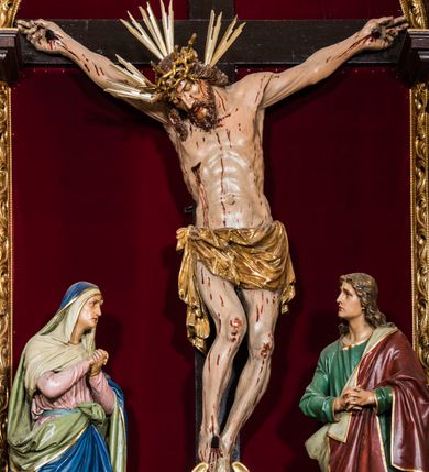 Zdjęcie nr 32: Ołtarz drewniany, architektoniczny, trójosiowy, jednokondygnacyjny ze zwieńczeniem usytuowany w połowie prezbiterium, na całej jego szerokości. Ołtarz prostopadłościenny z drewnianą mensą. Tabernakulum jest trójosiowe, osadzone na podwójnym cokole, zryzalitowane w części środkowej, podzielone kompozytowymi kolumnami, podtrzymującymi wyłamujące się nad nimi belkowanie z przerwanym przyczółkiem pośrodku. W części środkowej ruchomy tron wystawienia, wieloboczny, z rytymi literami IHS na tle glorii promienistej w jednej części i gałązkami z kwiatami w drugiej. Retabulum osadzone na wysokim cokole, z bramkami po bokach zamkniętymi łukami dwuramiennymi. Na dwiczkach malowane wizerunki św. Piotra po prawej i św. Pawła po lewej stronie. Część środkową retabulum ujmują dwie pary kompozytowych pilastrów o kanelowanych trzonech, z których środkowe cofnięte, podtrzymują przełamujące się nad nimi belkowanie i przerwany przyczółek. Zwieńczenie jest ujęte kanelowanymi pilastrami kompozytowymi z naczółkami oraz para ażurowych, ornamentacyjnych uszaków. W części środkowej jest wnęka w kształcie pionowego prostokąta zamkniętego łukiem półkolistym z rzeźbiarską grupą Ukrzyżowania z Matką Boską i św. Janem. W zwieńczeniu jest obraz „Pietas Domini&quot; w sześciobocznej ramie. W osiach bocznych nastawy znajdują się ażurowe arkady z malowanymi wizerunkami Chrystusa u słupa i Ecce Homo. Osie zwieńczone są półkolistymi przyczółkami. W osiach bocznych przeszklone galoty z kompozycją wykonaną z tkanin oraz srebrnych i złotych nici. Dekorację ornamentalną ołtarza i tabernakulum stanowią liście akantu, girlandy i rozety. Struktura utrzymana jest w kolorze brązowym, ornamentyka i detal architektoniczy są złocone. 