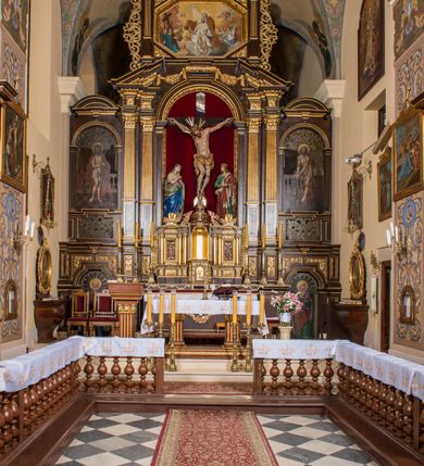 Zdjęcie nr 2: Ołtarz drewniany, architektoniczny, trójosiowy, jednokondygnacyjny ze zwieńczeniem usytuowany w połowie prezbiterium, na całej jego szerokości. Ołtarz prostopadłościenny z drewnianą mensą. Tabernakulum jest trójosiowe, osadzone na podwójnym cokole, zryzalitowane w części środkowej, podzielone kompozytowymi kolumnami, podtrzymującymi wyłamujące się nad nimi belkowanie z przerwanym przyczółkiem pośrodku. W części środkowej ruchomy tron wystawienia, wieloboczny, z rytymi literami IHS na tle glorii promienistej w jednej części i gałązkami z kwiatami w drugiej. Retabulum osadzone na wysokim cokole, z bramkami po bokach zamkniętymi łukami dwuramiennymi. Na dwiczkach malowane wizerunki św. Piotra po prawej i św. Pawła po lewej stronie. Część środkową retabulum ujmują dwie pary kompozytowych pilastrów o kanelowanych trzonech, z których środkowe cofnięte, podtrzymują przełamujące się nad nimi belkowanie i przerwany przyczółek. Zwieńczenie jest ujęte kanelowanymi pilastrami kompozytowymi z naczółkami oraz para ażurowych, ornamentacyjnych uszaków. W części środkowej jest wnęka w kształcie pionowego prostokąta zamkniętego łukiem półkolistym z rzeźbiarską grupą Ukrzyżowania z Matką Boską i św. Janem. W zwieńczeniu jest obraz „Pietas Domini&quot; w sześciobocznej ramie. W osiach bocznych nastawy znajdują się ażurowe arkady z malowanymi wizerunkami Chrystusa u słupa i Ecce Homo. Osie zwieńczone są półkolistymi przyczółkami. W osiach bocznych przeszklone galoty z kompozycją wykonaną z tkanin oraz srebrnych i złotych nici. Dekorację ornamentalną ołtarza i tabernakulum stanowią liście akantu, girlandy i rozety. Struktura utrzymana jest w kolorze brązowym, ornamentyka i detal architektoniczy są złocone. 