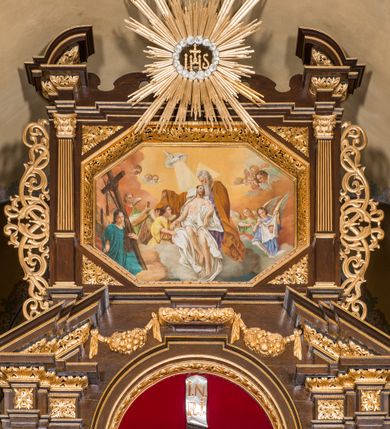 Zdjęcie nr 34: Ołtarz drewniany, architektoniczny, trójosiowy, jednokondygnacyjny ze zwieńczeniem usytuowany w połowie prezbiterium, na całej jego szerokości. Ołtarz prostopadłościenny z drewnianą mensą. Tabernakulum jest trójosiowe, osadzone na podwójnym cokole, zryzalitowane w części środkowej, podzielone kompozytowymi kolumnami, podtrzymującymi wyłamujące się nad nimi belkowanie z przerwanym przyczółkiem pośrodku. W części środkowej ruchomy tron wystawienia, wieloboczny, z rytymi literami IHS na tle glorii promienistej w jednej części i gałązkami z kwiatami w drugiej. Retabulum osadzone na wysokim cokole, z bramkami po bokach zamkniętymi łukami dwuramiennymi. Na dwiczkach malowane wizerunki św. Piotra po prawej i św. Pawła po lewej stronie. Część środkową retabulum ujmują dwie pary kompozytowych pilastrów o kanelowanych trzonech, z których środkowe cofnięte, podtrzymują przełamujące się nad nimi belkowanie i przerwany przyczółek. Zwieńczenie jest ujęte kanelowanymi pilastrami kompozytowymi z naczółkami oraz para ażurowych, ornamentacyjnych uszaków. W części środkowej jest wnęka w kształcie pionowego prostokąta zamkniętego łukiem półkolistym z rzeźbiarską grupą Ukrzyżowania z Matką Boską i św. Janem. W zwieńczeniu jest obraz „Pietas Domini" w sześciobocznej ramie. W osiach bocznych nastawy znajdują się ażurowe arkady z malowanymi wizerunkami Chrystusa u słupa i Ecce Homo. Osie zwieńczone są półkolistymi przyczółkami. W osiach bocznych przeszklone galoty z kompozycją wykonaną z tkanin oraz srebrnych i złotych nici. Dekorację ornamentalną ołtarza i tabernakulum stanowią liście akantu, girlandy i rozety. Struktura utrzymana jest w kolorze brązowym, ornamentyka i detal architektoniczy są złocone. 