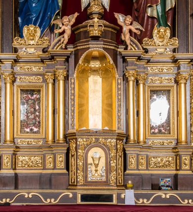 Zdjęcie nr 5: Ołtarz drewniany, architektoniczny, trójosiowy, jednokondygnacyjny ze zwieńczeniem usytuowany w połowie prezbiterium, na całej jego szerokości. Ołtarz prostopadłościenny z drewnianą mensą. Tabernakulum jest trójosiowe, osadzone na podwójnym cokole, zryzalitowane w części środkowej, podzielone kompozytowymi kolumnami, podtrzymującymi wyłamujące się nad nimi belkowanie z przerwanym przyczółkiem pośrodku. W części środkowej ruchomy tron wystawienia, wieloboczny, z rytymi literami IHS na tle glorii promienistej w jednej części i gałązkami z kwiatami w drugiej. Retabulum osadzone na wysokim cokole, z bramkami po bokach zamkniętymi łukami dwuramiennymi. Na dwiczkach malowane wizerunki św. Piotra po prawej i św. Pawła po lewej stronie. Część środkową retabulum ujmują dwie pary kompozytowych pilastrów o kanelowanych trzonech, z których środkowe cofnięte, podtrzymują przełamujące się nad nimi belkowanie i przerwany przyczółek. Zwieńczenie jest ujęte kanelowanymi pilastrami kompozytowymi z naczółkami oraz para ażurowych, ornamentacyjnych uszaków. W części środkowej jest wnęka w kształcie pionowego prostokąta zamkniętego łukiem półkolistym z rzeźbiarską grupą Ukrzyżowania z Matką Boską i św. Janem. W zwieńczeniu jest obraz „Pietas Domini" w sześciobocznej ramie. W osiach bocznych nastawy znajdują się ażurowe arkady z malowanymi wizerunkami Chrystusa u słupa i Ecce Homo. Osie zwieńczone są półkolistymi przyczółkami. W osiach bocznych przeszklone galoty z kompozycją wykonaną z tkanin oraz srebrnych i złotych nici. Dekorację ornamentalną ołtarza i tabernakulum stanowią liście akantu, girlandy i rozety. Struktura utrzymana jest w kolorze brązowym, ornamentyka i detal architektoniczy są złocone. 