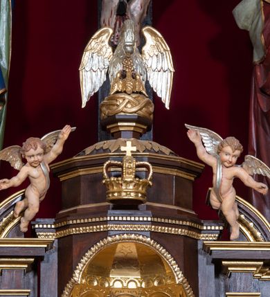 Zdjęcie nr 28: Ołtarz drewniany, architektoniczny, trójosiowy, jednokondygnacyjny ze zwieńczeniem usytuowany w połowie prezbiterium, na całej jego szerokości. Ołtarz prostopadłościenny z drewnianą mensą. Tabernakulum jest trójosiowe, osadzone na podwójnym cokole, zryzalitowane w części środkowej, podzielone kompozytowymi kolumnami, podtrzymującymi wyłamujące się nad nimi belkowanie z przerwanym przyczółkiem pośrodku. W części środkowej ruchomy tron wystawienia, wieloboczny, z rytymi literami IHS na tle glorii promienistej w jednej części i gałązkami z kwiatami w drugiej. Retabulum osadzone na wysokim cokole, z bramkami po bokach zamkniętymi łukami dwuramiennymi. Na dwiczkach malowane wizerunki św. Piotra po prawej i św. Pawła po lewej stronie. Część środkową retabulum ujmują dwie pary kompozytowych pilastrów o kanelowanych trzonech, z których środkowe cofnięte, podtrzymują przełamujące się nad nimi belkowanie i przerwany przyczółek. Zwieńczenie jest ujęte kanelowanymi pilastrami kompozytowymi z naczółkami oraz para ażurowych, ornamentacyjnych uszaków. W części środkowej jest wnęka w kształcie pionowego prostokąta zamkniętego łukiem półkolistym z rzeźbiarską grupą Ukrzyżowania z Matką Boską i św. Janem. W zwieńczeniu jest obraz „Pietas Domini" w sześciobocznej ramie. W osiach bocznych nastawy znajdują się ażurowe arkady z malowanymi wizerunkami Chrystusa u słupa i Ecce Homo. Osie zwieńczone są półkolistymi przyczółkami. W osiach bocznych przeszklone galoty z kompozycją wykonaną z tkanin oraz srebrnych i złotych nici. Dekorację ornamentalną ołtarza i tabernakulum stanowią liście akantu, girlandy i rozety. Struktura utrzymana jest w kolorze brązowym, ornamentyka i detal architektoniczy są złocone. 