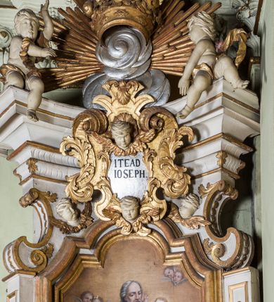 Zdjęcie nr 16: Ołtarz architektoniczny, jednokondygnacyjny, jednoosiowy, ze zwieńczeniem. Ołtarz prostopadłościenny, murowany. Retabulum o strukturze wklęsłej wzdłuż osi pionowej osadzone na niewysokim, cokole zryzalitowanym w formie pary impostów. Gzyms cokołu pośrodku ma formę półkolistego łuku nadwieszonego. Część środkowa ujęta parą wąskich filarów, licami zwróconych ukośnie w prawo, wspierających wydatny gzyms osadzony na parze spływów wolutowych. Pola cokołu retabulum zdobione ornamentyką roślinną. Na impostach para figur aniołków, stojący po lewej stronie trzyma w dłoniach zwój z napisem „KOCHAJ BOGA / I BLIZNIEGO”, stojący po prawej lewą rękę wspiera na kartuszu z napisem „POZNAJ / SIEBIE / SAMEGO”. Miedzy impostami płycina w kształcie prostokąta leżącego o wklęsłych górnych narożach i zamkniętego półkolistym łukiem nadwieszonym. W dolnej partii retabulum para uskrzydlonych główek anielskich dołem ujętych liśćmi akantu. Lica filarów zdobione motywem kampanuli. W partii belkowania, pośrodku motyw akantowego kartusza z napisem „ITE AD / IOSEPH.”, ujętego górą, dołem i po bokach motywami uskrzydlonych główek anielskich. Zwieńczenie w formie promienistej glorii z koroną zdobioną kwiatami osadzoną na obłokach. Całość flankuje para siedzących na gzymsie aniołów w dynamicznych pozach. Pole części środkowej w kształcie stojącego prostokąta ujętego górą i dołem parą zwróconych do siebie półkolistych łuków nadwieszonych z uskokami, ujęte profilowaną, złoconą ramą. Wewnątrz obraz z wizerunkiem św. Józefa. Struktura polichromowana w kolorze białym, detal architektoniczny i ornamentyka złocone i srebrzone.