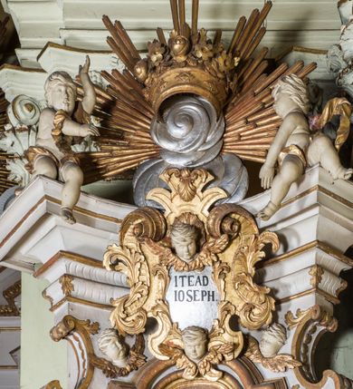 Zdjęcie nr 17: Ołtarz architektoniczny, jednokondygnacyjny, jednoosiowy, ze zwieńczeniem. Ołtarz prostopadłościenny, murowany. Retabulum o strukturze wklęsłej wzdłuż osi pionowej osadzone na niewysokim, cokole zryzalitowanym w formie pary impostów. Gzyms cokołu pośrodku ma formę półkolistego łuku nadwieszonego. Część środkowa ujęta parą wąskich filarów, licami zwróconych ukośnie w prawo, wspierających wydatny gzyms osadzony na parze spływów wolutowych. Pola cokołu retabulum zdobione ornamentyką roślinną. Na impostach para figur aniołków, stojący po lewej stronie trzyma w dłoniach zwój z napisem „KOCHAJ BOGA / I BLIZNIEGO”, stojący po prawej lewą rękę wspiera na kartuszu z napisem „POZNAJ / SIEBIE / SAMEGO”. Miedzy impostami płycina w kształcie prostokąta leżącego o wklęsłych górnych narożach i zamkniętego półkolistym łukiem nadwieszonym. W dolnej partii retabulum para uskrzydlonych główek anielskich dołem ujętych liśćmi akantu. Lica filarów zdobione motywem kampanuli. W partii belkowania, pośrodku motyw akantowego kartusza z napisem „ITE AD / IOSEPH.”, ujętego górą, dołem i po bokach motywami uskrzydlonych główek anielskich. Zwieńczenie w formie promienistej glorii z koroną zdobioną kwiatami osadzoną na obłokach. Całość flankuje para siedzących na gzymsie aniołów w dynamicznych pozach. Pole części środkowej w kształcie stojącego prostokąta ujętego górą i dołem parą zwróconych do siebie półkolistych łuków nadwieszonych z uskokami, ujęte profilowaną, złoconą ramą. Wewnątrz obraz z wizerunkiem św. Józefa. Struktura polichromowana w kolorze białym, detal architektoniczny i ornamentyka złocone i srebrzone.