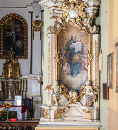 Zdjęcie nr 2: Ołtarz architektoniczny, jednokondygnacyjny, jednoosiowy, ze zwieńczeniem. Ołtarz prostopadłościenny, murowany. Retabulum o strukturze wklęsłej wzdłuż osi pionowej osadzone na niewysokim, cokole zryzalitowanym w formie pary impostów. Gzyms cokołu pośrodku ma formę półkolistego łuku nadwieszonego. Część środkowa ujęta parą wąskich filarów, licami zwróconych ukośnie w prawo, wspierających wydatny gzyms osadzony na parze spływów wolutowych. Pola cokołu retabulum zdobione ornamentyką roślinną. Na impostach para figur aniołków, stojący po lewej stronie trzyma w dłoniach zwój z napisem „KOCHAJ BOGA / I BLIZNIEGO”, stojący po prawej lewą rękę wspiera na kartuszu z napisem „POZNAJ / SIEBIE / SAMEGO”. Miedzy impostami płycina w kształcie prostokąta leżącego o wklęsłych górnych narożach i zamkniętego półkolistym łukiem nadwieszonym. W dolnej partii retabulum para uskrzydlonych główek anielskich dołem ujętych liśćmi akantu. Lica filarów zdobione motywem kampanuli. W partii belkowania, pośrodku motyw akantowego kartusza z napisem „ITE AD / IOSEPH.”, ujętego górą, dołem i po bokach motywami uskrzydlonych główek anielskich. Zwieńczenie w formie promienistej glorii z koroną zdobioną kwiatami osadzoną na obłokach. Całość flankuje para siedzących na gzymsie aniołów w dynamicznych pozach. Pole części środkowej w kształcie stojącego prostokąta ujętego górą i dołem parą zwróconych do siebie półkolistych łuków nadwieszonych z uskokami, ujęte profilowaną, złoconą ramą. Wewnątrz obraz z wizerunkiem św. Józefa. Struktura polichromowana w kolorze białym, detal architektoniczny i ornamentyka złocone i srebrzone.