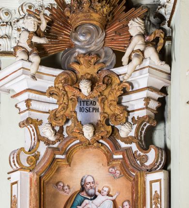 Zdjęcie nr 14: Ołtarz architektoniczny, jednokondygnacyjny, jednoosiowy, ze zwieńczeniem. Ołtarz prostopadłościenny, murowany. Retabulum o strukturze wklęsłej wzdłuż osi pionowej osadzone na niewysokim, cokole zryzalitowanym w formie pary impostów. Gzyms cokołu pośrodku ma formę półkolistego łuku nadwieszonego. Część środkowa ujęta parą wąskich filarów, licami zwróconych ukośnie w prawo, wspierających wydatny gzyms osadzony na parze spływów wolutowych. Pola cokołu retabulum zdobione ornamentyką roślinną. Na impostach para figur aniołków, stojący po lewej stronie trzyma w dłoniach zwój z napisem „KOCHAJ BOGA / I BLIZNIEGO”, stojący po prawej lewą rękę wspiera na kartuszu z napisem „POZNAJ / SIEBIE / SAMEGO”. Miedzy impostami płycina w kształcie prostokąta leżącego o wklęsłych górnych narożach i zamkniętego półkolistym łukiem nadwieszonym. W dolnej partii retabulum para uskrzydlonych główek anielskich dołem ujętych liśćmi akantu. Lica filarów zdobione motywem kampanuli. W partii belkowania, pośrodku motyw akantowego kartusza z napisem „ITE AD / IOSEPH.”, ujętego górą, dołem i po bokach motywami uskrzydlonych główek anielskich. Zwieńczenie w formie promienistej glorii z koroną zdobioną kwiatami osadzoną na obłokach. Całość flankuje para siedzących na gzymsie aniołów w dynamicznych pozach. Pole części środkowej w kształcie stojącego prostokąta ujętego górą i dołem parą zwróconych do siebie półkolistych łuków nadwieszonych z uskokami, ujęte profilowaną, złoconą ramą. Wewnątrz obraz z wizerunkiem św. Józefa. Struktura polichromowana w kolorze białym, detal architektoniczny i ornamentyka złocone i srebrzone.