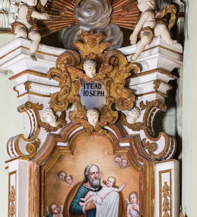 Zdjęcie nr 15: Ołtarz architektoniczny, jednokondygnacyjny, jednoosiowy, ze zwieńczeniem. Ołtarz prostopadłościenny, murowany. Retabulum o strukturze wklęsłej wzdłuż osi pionowej osadzone na niewysokim, cokole zryzalitowanym w formie pary impostów. Gzyms cokołu pośrodku ma formę półkolistego łuku nadwieszonego. Część środkowa ujęta parą wąskich filarów, licami zwróconych ukośnie w prawo, wspierających wydatny gzyms osadzony na parze spływów wolutowych. Pola cokołu retabulum zdobione ornamentyką roślinną. Na impostach para figur aniołków, stojący po lewej stronie trzyma w dłoniach zwój z napisem „KOCHAJ BOGA / I BLIZNIEGO”, stojący po prawej lewą rękę wspiera na kartuszu z napisem „POZNAJ / SIEBIE / SAMEGO”. Miedzy impostami płycina w kształcie prostokąta leżącego o wklęsłych górnych narożach i zamkniętego półkolistym łukiem nadwieszonym. W dolnej partii retabulum para uskrzydlonych główek anielskich dołem ujętych liśćmi akantu. Lica filarów zdobione motywem kampanuli. W partii belkowania, pośrodku motyw akantowego kartusza z napisem „ITE AD / IOSEPH.”, ujętego górą, dołem i po bokach motywami uskrzydlonych główek anielskich. Zwieńczenie w formie promienistej glorii z koroną zdobioną kwiatami osadzoną na obłokach. Całość flankuje para siedzących na gzymsie aniołów w dynamicznych pozach. Pole części środkowej w kształcie stojącego prostokąta ujętego górą i dołem parą zwróconych do siebie półkolistych łuków nadwieszonych z uskokami, ujęte profilowaną, złoconą ramą. Wewnątrz obraz z wizerunkiem św. Józefa. Struktura polichromowana w kolorze białym, detal architektoniczny i ornamentyka złocone i srebrzone.
