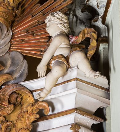 Zdjęcie nr 18: Ołtarz architektoniczny, jednokondygnacyjny, jednoosiowy, ze zwieńczeniem. Ołtarz prostopadłościenny, murowany. Retabulum o strukturze wklęsłej wzdłuż osi pionowej osadzone na niewysokim, cokole zryzalitowanym w formie pary impostów. Gzyms cokołu pośrodku ma formę półkolistego łuku nadwieszonego. Część środkowa ujęta parą wąskich filarów, licami zwróconych ukośnie w prawo, wspierających wydatny gzyms osadzony na parze spływów wolutowych. Pola cokołu retabulum zdobione ornamentyką roślinną. Na impostach para figur aniołków, stojący po lewej stronie trzyma w dłoniach zwój z napisem „KOCHAJ BOGA / I BLIZNIEGO”, stojący po prawej lewą rękę wspiera na kartuszu z napisem „POZNAJ / SIEBIE / SAMEGO”. Miedzy impostami płycina w kształcie prostokąta leżącego o wklęsłych górnych narożach i zamkniętego półkolistym łukiem nadwieszonym. W dolnej partii retabulum para uskrzydlonych główek anielskich dołem ujętych liśćmi akantu. Lica filarów zdobione motywem kampanuli. W partii belkowania, pośrodku motyw akantowego kartusza z napisem „ITE AD / IOSEPH.”, ujętego górą, dołem i po bokach motywami uskrzydlonych główek anielskich. Zwieńczenie w formie promienistej glorii z koroną zdobioną kwiatami osadzoną na obłokach. Całość flankuje para siedzących na gzymsie aniołów w dynamicznych pozach. Pole części środkowej w kształcie stojącego prostokąta ujętego górą i dołem parą zwróconych do siebie półkolistych łuków nadwieszonych z uskokami, ujęte profilowaną, złoconą ramą. Wewnątrz obraz z wizerunkiem św. Józefa. Struktura polichromowana w kolorze białym, detal architektoniczny i ornamentyka złocone i srebrzone.