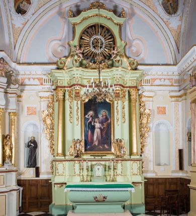 Zdjęcie nr 1: Ołtarz architektoniczny, jednokondygnacyjny, jednoosiowy, ze zwieńczeniem. Ołtarz sarkofagowy, z drewnianą mensą, pośrodku pola antepedium polichromowany monogram maryjny ujęty parą gałązek różanych. Tabernakulum w formie prostopadłościennej szafki osadzonej na zdwojonym cokole, o narożach ujętych parą skośnie ustawionych pilastrów wspierających wyłamujący się ponad nimi gzyms wieńczący, w części środkowej przybierający formę dwóch stykających się wolut. Drzwiczki są w kształcie stojącego prostokąta zamkniętego łukiem półkolistym, ujęte profilowaną, złoconą ramą. W polu płaskorzeźbiony motyw kielicha eucharystycznego. Retabulum osadzone na wysokim, zdwojonym cokole. Część środkowa ujęta parą filarów i parą cofniętych kolumn o kapitelach korynckich. Podpory wspierają wyłamujące się ponad nimi belkowanie z wydatnym gzymsem. Zwieńczenie w formie trapezoidalnej ujęte parą spływów wolutowych, zamknięte fragmentem gzymsu w postaci nawieszonego łuku odcinkowego z uskokami, dodatkowo flankowane przerwanym przyczółkiem wolutowym. Dolny cokół retabulum zdobiony motywami kogucich grzebieni, pola górnego artykułowane fragmentami ornamentu roślinnego, całość ujęta małymi uszakami skomponowanymi z kogucich grzebieni. Trzony filarów dołem zdobione rocaille’owymi kartuszami, górą podwieszenia rocaille’owe. Fryz zdobiony motywami ornamentyki roślinnej. Retabulum ujęte parą ażurowych uszaków skomponowanych z wici roślinnej i kogucich grzebieni. W zwieńczeniu, pośrodku wsparta na belkowaniu duża korona, na przerwanym przyczółku para aniołków z ulistnionymi gałązkami w podniesionych rękach. Gzyms wieńczący ujęty dołem rodzajem lambrekinu z pojedynczych liści akantu. Szczyt w formie ażurowego kartusza rocaille’owego ujęty po bokach parą płomienistych wazonów. Pole części środkowej w kształcie stojącego prostokąta zamkniętego półkolistym łukiem nawieszonym z uskokami, ujęte profilowaną, złoconą ramą. W środku obraz „Święta Rodzina” („Znalezienie Jezusa w świątyni”), na cokole retabulum para figur, po lewej św. Michał Archanioł, po prawej św. Rafał Archanioł. Pole zwieńczenia w postaci otworu w kształcie stojącego owalu wypełnione ażurową, promienistą glorią z monogramem maryjnym pośrodku ujętym wieńcem obłoków. Struktura polichromowana w odcieniu jasnej zieleni, detal architektoniczny i ornamentyka polichromowane w kolorze bordowym, złocone i srebrzone. 