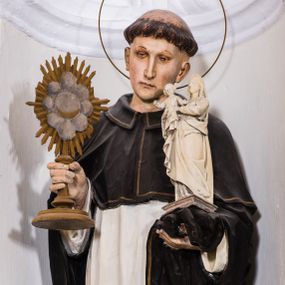 Zdjęcie nr 1: Całopostaciowa, pełnoplastyczna figura przedstawia zwróconego na wprost św. Jacka Odrowąża stojącego w lekkim kontrapoście na niewysokiej, prostopadłościennej podstawie o ściętych narożach. Lekko przechyloną w lewo głowę świętego charakteryzuje smukła, pociągła twarz o delikatnych rysach z długim, prostym nosem, skierowanym w dół wzrokiem i zarumienionymi policzkami. Krótkie, brązowe włosy przycięte są w formie tonsury. Święty ubrany jest w habit dominikański z białą stułą na ramionach i czarną kukullą. Na stopach ma czarne buty, głowę okala wąska aureola. W prawej ręce trzyma złoconą monstrancję, w lewej, na pole płaszcza podtrzymuje niewielki, osadzony na niewysokim, profilowanym postumencie, biały posążek Madonny trzymającej oburącz Dzieciątko Jezus. 