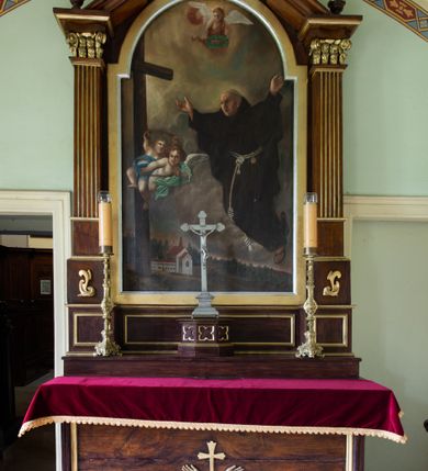 Zdjęcie nr 2: Obraz w kształcie stojącego prostokąta zamkniętego półkolistym łukiem nadwieszonym przedstawia św. Piotra z Alkantary. Zajmująca prawą stronę kompozycji, zwrócona w trzech czwartych w lewo sylwetka świętego unosi się w powietrzu. Twarz mężczyzny charakteryzuje długi, spiczasty nos, lekko rozchylone usta i dwie bruzdy na czole między oczami. Włosy przystrzyżone są w formie tonsury. Święty ubrany jest w habit franciszkański, na stopach ma sandały. Rozłożone na boki ręce wznosi do góry. Wzrok kieruje w stronę znajdującego się po lewej stronie kompozycji krzyża, z którego biją jasne, delikatne promienie. W połowie wysokości pionowej belki unosi się w powietrzu para aniołków częściowo przesłoniętych niebieską i zieloną szatą, trzymających w dłoniach łańcuch i pas nabity gwoźdźmi. Trzeci anioł, ubrany w czerwoną szatę, ukazany jest w górnej partii kompozycji, oburącz podtrzymując w dłoniach wieniec laurowy. Postaci ukazane są na tle pejzażu o obniżonej linii horyzontu, którego znaczną część stanowi pokryte gęstymi chmurami niebo, w dole zaś widoczna jest zabudowa klasztorna oraz las. W lewym, dolnym rogu płótna znajduje się sygnatura „Pinx(it) A. Kowarz / Reform. 1873.”.    