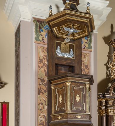 Zdjęcie nr 2: Ambona przyścienna, wisząca, z wejściem od strony zaplecka. Kosz jest na planie ściętego sześcioboku ze ściankami wydzielonymi półfilarami o kapitelach korynckich. Pola ścianek wypełniają płyciny w kształcie stojącego prostokąta o narożach w formie uszaków, ujęte profilowanym obramieniem. Dół kosza obiega belkowanie z wybrzuszonym architrawem, szerokim fryzem i delikatnym gzymsem. Podwieszenie kosza zakończone jest motywem złoconej szyszki. Parapet ambony jest w formie belkowania ze znacznie wysuniętym gzymsem. W dolnej części zaplecka znajdują się prowadzące na ambonę drzwi. Powyżej jest płycina w kształcie leżącego prostokąta z płaskorzeźbioną, złoconą parą tablic Mojżeszowych, ujętych srebrzonymi obłokami. Baldachim na rzucie sześcioboku tworzy sześć wklęsłych płycin, dołem obiega je belkowanie z wydatnym gzymsem. Powyżej gzymsu znajduje się niewysoki cokół wyłamujący się na narożach, na których ustawione są płonące wazony. Baldachim wieńczy krzyż. Płyciny kosza ambony zdobią motywy stylizowanej ornamentyki roślinnej, w podniebiu znajduje się sylwetka gołębicy Ducha Świętego ujętej promienistą glorią. Struktura ambony utrzymana jest w odcieniach brązu, detal architektoniczny i ornamentyka są złocone i srebrzone.   