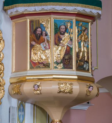 Zdjęcie nr 2: Wisząca, przyścienna ambona składa się z kosza, zaplecka i baldachimu. Dojście do kosza prowadzi przez dwuskrzydłowe drzwi usytuowane w zaplecku. Kształt kosza oparto na planie siedmiu boków z dziesięcioboku, ustawionych na wałku, z podwieszeniem w formie zbliżonej do stożka, zakończonego szyszką oraz dekorowanym rozetami i uskrzydlonymi główkami anielskimi. Ścianki kosza artykułowano prostokątnymi, obramienionymi płycinami z pięcioma płaskorzeźbionymi przedstawieniam czterech ewangelistów i siewcy. Każdego ewangelistę identyfikuje jego atrybut. Siewca, umieszczony na trzeciej płycinie, został ukazany jako młody mężczyzna, który idąc przez zaorane pole rzuca w nie ziarno. U jego stóp widać pług oraz worek ze zbożem na zasiew. Parapet kosza uzyskał formę gzymsu. Zaplecek został ujęty profilowaną ramą. W jego pole wpisano prostokątne, dwuskrzydłowe drzwi w profilowanym obramieniu. Baldachim uzyskał rzut analogiczny do kosza ambony. Płaskie podniebie z wyobrażeniem gołębicy Ducha Świętego na tle złocistych promieni ujmuje profilowany, łamany gzyms, ponad którym poprowadzono kopulasty dach. Na jego szczycie ustawiono rzeźbę Mojżesza trzymającego tablice Dekalogu. Struktura ambony została polichromowana w kolorze beżowym, z białymi i czerwonymi profilami, oraz pozłacanymi detalami, profilami i ornamentyką. Płaskorzeźby na koszu oraz figura Mojżesza otrzymały naturalistyczną polichromię.