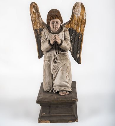 Zdjęcie nr 1: Pełnoplastyczna figura ukazuje przyklękającego na prawe kolano uskrzydlonego anioła. Anioł o lekko pochylonej głowie do przodu ma modlitewnie złożone ręce przed sobą. Jego owalną twarz o jasnej karnacji, drobnym nosie i ustach otaczają krótkie brązowe włosy. Postać ubrana jest w białą, luźną tunikę. Umieszono ją na prostopadłościennym drewnianym postumencie zaopatrzonym w niski profilowany cokół oraz gzyms. 