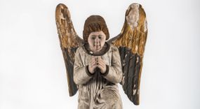 Zdjęcie nr 1: Pełnoplastyczna figura ukazuje przyklękającego na prawe kolano uskrzydlonego anioła. Anioł o lekko pochylonej głowie do przodu ma modlitewnie złożone ręce przed sobą. Jego owalną twarz o jasnej karnacji, drobnym nosie i ustach otaczają krótkie brązowe włosy. Postać ubrana jest w białą, luźną tunikę. Umieszono ją na prostopadłościennym drewnianym postumencie zaopatrzonym w niski profilowany cokół oraz gzyms. 