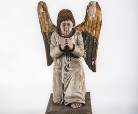 Zdjęcie nr 1: Pełnoplastyczna figura ukazuje przyklękającego na prawe kolano uskrzydlonego anioła. Anioł o lekko pochylonej głowie do przodu ma modlitewnie złożone ręce przed sobą. Jego owalną twarz o jasnej karnacji, drobnym nosie i ustach otaczają krótkie brązowe włosy. Postać ubrana jest w białą, luźną tunikę. Umieszono ją na prostopadłościennym drewnianym postumencie zaopatrzonym w niski profilowany cokół oraz gzyms. 