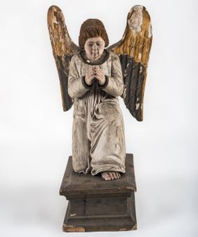 Zdjęcie nr 1: Pełnoplastyczna figura ukazuje przyklękającego na prawe kolano uskrzydlonego anioła. Anioł o lekko pochylonej głowie do przodu ma modlitewnie złożone ręce przed sobą. Jego owalną twarz o jasnej karnacji, drobnym nosie i ustach otaczają krótkie brązowe włosy. Postać ubrana jest w białą, luźną tunikę. Umieszono ją na prostopadłościennym drewnianym postumencie zaopatrzonym w niski profilowany cokół oraz gzyms. 