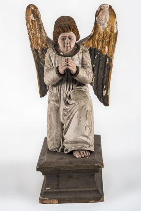 Zdjęcie nr 1: Pełnoplastyczna figura ukazuje przyklękającego na prawe kolano uskrzydlonego anioła. Anioł o lekko pochylonej głowie do przodu ma modlitewnie złożone ręce przed sobą. Jego owalną twarz o jasnej karnacji, drobnym nosie i ustach otaczają krótkie brązowe włosy. Postać ubrana jest w białą, luźną tunikę. Umieszono ją na prostopadłościennym drewnianym postumencie zaopatrzonym w niski profilowany cokół oraz gzyms. 
