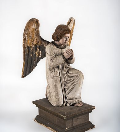 Zdjęcie nr 2: Pełnoplastyczna figura ukazuje przyklękającego na prawe kolano uskrzydlonego anioła. Anioł o lekko pochylonej głowie do przodu ma modlitewnie złożone ręce przed sobą. Jego owalną twarz o jasnej karnacji, drobnym nosie i ustach otaczają krótkie brązowe włosy. Postać ubrana jest w białą, luźną tunikę. Umieszono ją na prostopadłościennym drewnianym postumencie zaopatrzonym w niski profilowany cokół oraz gzyms. 