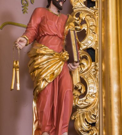 Zdjęcie nr 3: Rzeźba św. Piotra została ścięta i wydrążona z tyłu oraz ustawiona na podstawie na planie kwadratu. Święty został ujęty frontalnie, w kontrapoście. Lewą ręką podtrzymuje przy boku zamknietą księgę, a w prawej wyciągniętej w bok trzyma dwa klucze. Apostoł ma pociągłą twarz o wyrazistych rysach, głęboko osadzonych oczach, prostym nosie i lekko rozchylonych ustach. Głowę okala krótka brodą i krótkie, kręcone, ciemne włosy z charkaterystycznym puklem na czole. Ubrany jest w różowo-pomarańczową tunikę ze złoconą lamówką oraz złocony płaszcz przewiązany diagonalnie przez lewe ramię i zawiązany z przodu postaci. Spod tuniki widać bose stopy. Polichromia ciała jest naturalistyczna.