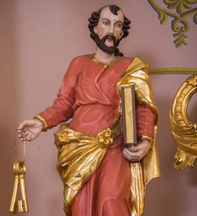 Zdjęcie nr 1: Rzeźba św. Piotra została ścięta i wydrążona z tyłu oraz ustawiona na podstawie na planie kwadratu. Święty został ujęty frontalnie, w kontrapoście. Lewą ręką podtrzymuje przy boku zamknietą księgę, a w prawej wyciągniętej w bok trzyma dwa klucze. Apostoł ma pociągłą twarz o wyrazistych rysach, głęboko osadzonych oczach, prostym nosie i lekko rozchylonych ustach. Głowę okala krótka brodą i krótkie, kręcone, ciemne włosy z charkaterystycznym puklem na czole. Ubrany jest w różowo-pomarańczową tunikę ze złoconą lamówką oraz złocony płaszcz przewiązany diagonalnie przez lewe ramię i zawiązany z przodu postaci. Spod tuniki widać bose stopy. Polichromia ciała jest naturalistyczna.