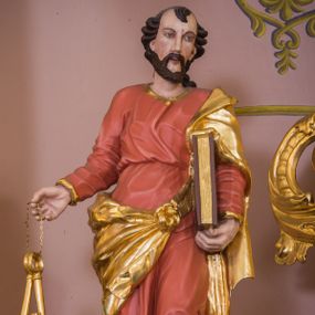 Zdjęcie nr 1: Rzeźba św. Piotra została ścięta i wydrążona z tyłu oraz ustawiona na podstawie na planie kwadratu. Święty został ujęty frontalnie, w kontrapoście. Lewą ręką podtrzymuje przy boku zamknietą księgę, a w prawej wyciągniętej w bok trzyma dwa klucze. Apostoł ma pociągłą twarz o wyrazistych rysach, głęboko osadzonych oczach, prostym nosie i lekko rozchylonych ustach. Głowę okala krótka brodą i krótkie, kręcone, ciemne włosy z charkaterystycznym puklem na czole. Ubrany jest w różowo-pomarańczową tunikę ze złoconą lamówką oraz złocony płaszcz przewiązany diagonalnie przez lewe ramię i zawiązany z przodu postaci. Spod tuniki widać bose stopy. Polichromia ciała jest naturalistyczna.