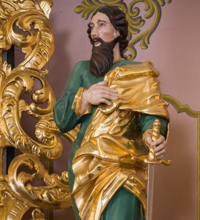 Zdjęcie nr 1: Rzeźba św. Pawła została ścięta z tyłu i wydrążona oraz ustawiona na kwadratowej podstawie. Apostoł został ukazany w trzech czwartych w lewo, w kontrapoście. W wyciągniętej w bok lewej ręce trzyma miecz zwrócony ostrzem w dół, prawą zaś wspiera na piersi. Twarz Pawła jest szczupła, o głęboko osadzonych oczach, prostym nosie i rozchylonych ustach. Okala ją długa skręcona broda opadająca na pierś oraz krótkie, kręcone włosy. Apostoł jest ubrany w zieloną tunikę, przylegająca do ciała o pozłacanych mankietach oraz płaszcz zawieszony na lewym ramieniu, opadający z przodu postaci i na prawy bok. Polichromia ciała jest naturalistyczna.