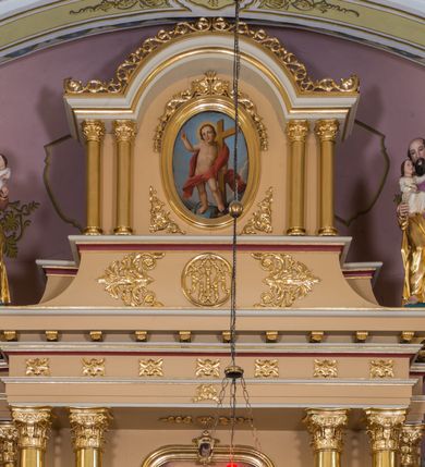 Zdjęcie nr 13: Jednokondygnacyjna nastawa ze zwieńczeniem uzyskała architektoniczną strukturę wzbogaconą o  flankujące ją bramkami. Współczesny marmurowy stipes ołtarzowy wspierający mensę uzyskał formę prostopadłościanu z licem artykułowanym dwiema półkolumienkami. Na mensie ustawiono tabernakulum w formie drewnianej skrzynki, obitej trybowaną blachą. Drzwiczki uzyskały kształt stojącego prostokąta zamkniętego łukiem półkolistym. W ich polu wyobrażono kielich z hostią w glorii, ujęty trybowanym winnym gronem i aplikowanymi kłosami zbóż. Z dwóch stron drzwiczek widnieje trybowana dekoracja w postaci winnego grona. Na tabernakulum usytuowano tron wystawienia, w postaci obrotowej niszy ujętej dwiema parami kolumienek podtrzymujących gzyms wyłamany pośrodku na kształt łuku odcinkowego oraz cokół z figurką baranka na księdze z siedmioma pieczęciami, podtrzymywany przez dwa spływy wolutowe. Tabernakulum ujęte zostało dwoma postumentami dekorowanymi z przodu rozetami. Ustawiono na nich figury klęczących aniołów. Nastawa ujęta została dwiema parami kolumn o kapitelach kompozytowych ustawionych na cokołach dekorowanych płycinami z ornamentem roślinnym, podtrzymujących pełne belkowanie z silnie wysuniętym gzymsem, oraz zewnętrzną parą cofniętych w tył kolumn. W polu głównym usytuowano obraz z przedstawieniem Zmartwychwstałego Chrystusa ze św. Janem Chrzcicielem i św. Janem Ewangelistą, ujęty profilowaną ramą w formie stojącego prostokąta zamkniętego łukiem odcinkowych nadwieszonym. Kondygnacja ujęta została po bokach uszami utworzonymi z karbowanej wstęgi i akantu. Zwieńczenie nastawy ustawiono na schodkowym, zwężającym się cokole, dekorowanym medalionem z monogramem Marii oraz anthemionami. Ujmują je dwie pary kolumienek o kapitelach kompozytowych podtrzymujących gzyms wyłamujący się po środku na kształt łuku półkolistego, dekorowany wicią roślinną. W polu zwieńczenia znalazł się obraz w formie stojącego owalu z przedstawieniem Dzieciątka Jezus prezentującego krzyż, ujęty profilowaną ramą dekorowaną ornamentem roślinnym. Zwieńczenie flankują stojące na gzymsie figury św. Józefa z Dzieciątkiem i Symeona z Dzieciątkiem. Nastawę flankują znajdujące się przy ścianie para kolumn i para półkolumn ustawionych na dwustrefowych cokołach, dźwigających pełne belkowanie, na którym siedzą dwa putta. Pomiędzy cokołem nastawy i zewnętrznych podpór ulokowano wygięte listwy tworzące bramki, na których stoją figury św. Piotra i św. Pawła. Struktura retabulum jest polichromowana w kolorze beżowym, z białymi i czerwonymi profilami i płycinami. Kolumny, ornamentyka i profile zostały pozłocone.