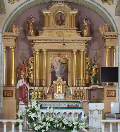 Zdjęcie nr 1: Jednokondygnacyjna nastawa ze zwieńczeniem uzyskała architektoniczną strukturę wzbogaconą o  flankujące ją bramkami. Współczesny marmurowy stipes ołtarzowy wspierający mensę uzyskał formę prostopadłościanu z licem artykułowanym dwiema półkolumienkami. Na mensie ustawiono tabernakulum w formie drewnianej skrzynki, obitej trybowaną blachą. Drzwiczki uzyskały kształt stojącego prostokąta zamkniętego łukiem półkolistym. W ich polu wyobrażono kielich z hostią w glorii, ujęty trybowanym winnym gronem i aplikowanymi kłosami zbóż. Z dwóch stron drzwiczek widnieje trybowana dekoracja w postaci winnego grona. Na tabernakulum usytuowano tron wystawienia, w postaci obrotowej niszy ujętej dwiema parami kolumienek podtrzymujących gzyms wyłamany pośrodku na kształt łuku odcinkowego oraz cokół z figurką baranka na księdze z siedmioma pieczęciami, podtrzymywany przez dwa spływy wolutowe. Tabernakulum ujęte zostało dwoma postumentami dekorowanymi z przodu rozetami. Ustawiono na nich figury klęczących aniołów. Nastawa ujęta została dwiema parami kolumn o kapitelach kompozytowych ustawionych na cokołach dekorowanych płycinami z ornamentem roślinnym, podtrzymujących pełne belkowanie z silnie wysuniętym gzymsem, oraz zewnętrzną parą cofniętych w tył kolumn. W polu głównym usytuowano obraz z przedstawieniem Zmartwychwstałego Chrystusa ze św. Janem Chrzcicielem i św. Janem Ewangelistą, ujęty profilowaną ramą w formie stojącego prostokąta zamkniętego łukiem odcinkowych nadwieszonym. Kondygnacja ujęta została po bokach uszami utworzonymi z karbowanej wstęgi i akantu. Zwieńczenie nastawy ustawiono na schodkowym, zwężającym się cokole, dekorowanym medalionem z monogramem Marii oraz anthemionami. Ujmują je dwie pary kolumienek o kapitelach kompozytowych podtrzymujących gzyms wyłamujący się po środku na kształt łuku półkolistego, dekorowany wicią roślinną. W polu zwieńczenia znalazł się obraz w formie stojącego owalu z przedstawieniem Dzieciątka Jezus prezentującego krzyż, ujęty profilowaną ramą dekorowaną ornamentem roślinnym. Zwieńczenie flankują stojące na gzymsie figury św. Józefa z Dzieciątkiem i Symeona z Dzieciątkiem. Nastawę flankują znajdujące się przy ścianie para kolumn i para półkolumn ustawionych na dwustrefowych cokołach, dźwigających pełne belkowanie, na którym siedzą dwa putta. Pomiędzy cokołem nastawy i zewnętrznych podpór ulokowano wygięte listwy tworzące bramki, na których stoją figury św. Piotra i św. Pawła. Struktura retabulum jest polichromowana w kolorze beżowym, z białymi i czerwonymi profilami i płycinami. Kolumny, ornamentyka i profile zostały pozłocone.