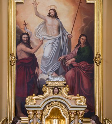 Zdjęcie nr 19: Jednokondygnacyjna nastawa ze zwieńczeniem uzyskała architektoniczną strukturę wzbogaconą o  flankujące ją bramkami. Współczesny marmurowy stipes ołtarzowy wspierający mensę uzyskał formę prostopadłościanu z licem artykułowanym dwiema półkolumienkami. Na mensie ustawiono tabernakulum w formie drewnianej skrzynki, obitej trybowaną blachą. Drzwiczki uzyskały kształt stojącego prostokąta zamkniętego łukiem półkolistym. W ich polu wyobrażono kielich z hostią w glorii, ujęty trybowanym winnym gronem i aplikowanymi kłosami zbóż. Z dwóch stron drzwiczek widnieje trybowana dekoracja w postaci winnego grona. Na tabernakulum usytuowano tron wystawienia, w postaci obrotowej niszy ujętej dwiema parami kolumienek podtrzymujących gzyms wyłamany pośrodku na kształt łuku odcinkowego oraz cokół z figurką baranka na księdze z siedmioma pieczęciami, podtrzymywany przez dwa spływy wolutowe. Tabernakulum ujęte zostało dwoma postumentami dekorowanymi z przodu rozetami. Ustawiono na nich figury klęczących aniołów. Nastawa ujęta została dwiema parami kolumn o kapitelach kompozytowych ustawionych na cokołach dekorowanych płycinami z ornamentem roślinnym, podtrzymujących pełne belkowanie z silnie wysuniętym gzymsem, oraz zewnętrzną parą cofniętych w tył kolumn. W polu głównym usytuowano obraz z przedstawieniem Zmartwychwstałego Chrystusa ze św. Janem Chrzcicielem i św. Janem Ewangelistą, ujęty profilowaną ramą w formie stojącego prostokąta zamkniętego łukiem odcinkowych nadwieszonym. Kondygnacja ujęta została po bokach uszami utworzonymi z karbowanej wstęgi i akantu. Zwieńczenie nastawy ustawiono na schodkowym, zwężającym się cokole, dekorowanym medalionem z monogramem Marii oraz anthemionami. Ujmują je dwie pary kolumienek o kapitelach kompozytowych podtrzymujących gzyms wyłamujący się po środku na kształt łuku półkolistego, dekorowany wicią roślinną. W polu zwieńczenia znalazł się obraz w formie stojącego owalu z przedstawieniem Dzieciątka Jezus prezentującego krzyż, ujęty profilowaną ramą dekorowaną ornamentem roślinnym. Zwieńczenie flankują stojące na gzymsie figury św. Józefa z Dzieciątkiem i Symeona z Dzieciątkiem. Nastawę flankują znajdujące się przy ścianie para kolumn i para półkolumn ustawionych na dwustrefowych cokołach, dźwigających pełne belkowanie, na którym siedzą dwa putta. Pomiędzy cokołem nastawy i zewnętrznych podpór ulokowano wygięte listwy tworzące bramki, na których stoją figury św. Piotra i św. Pawła. Struktura retabulum jest polichromowana w kolorze beżowym, z białymi i czerwonymi profilami i płycinami. Kolumny, ornamentyka i profile zostały pozłocone.