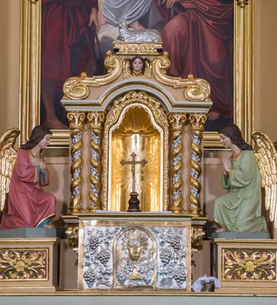 Zdjęcie nr 22: Jednokondygnacyjna nastawa ze zwieńczeniem uzyskała architektoniczną strukturę wzbogaconą o  flankujące ją bramkami. Współczesny marmurowy stipes ołtarzowy wspierający mensę uzyskał formę prostopadłościanu z licem artykułowanym dwiema półkolumienkami. Na mensie ustawiono tabernakulum w formie drewnianej skrzynki, obitej trybowaną blachą. Drzwiczki uzyskały kształt stojącego prostokąta zamkniętego łukiem półkolistym. W ich polu wyobrażono kielich z hostią w glorii, ujęty trybowanym winnym gronem i aplikowanymi kłosami zbóż. Z dwóch stron drzwiczek widnieje trybowana dekoracja w postaci winnego grona. Na tabernakulum usytuowano tron wystawienia, w postaci obrotowej niszy ujętej dwiema parami kolumienek podtrzymujących gzyms wyłamany pośrodku na kształt łuku odcinkowego oraz cokół z figurką baranka na księdze z siedmioma pieczęciami, podtrzymywany przez dwa spływy wolutowe. Tabernakulum ujęte zostało dwoma postumentami dekorowanymi z przodu rozetami. Ustawiono na nich figury klęczących aniołów. Nastawa ujęta została dwiema parami kolumn o kapitelach kompozytowych ustawionych na cokołach dekorowanych płycinami z ornamentem roślinnym, podtrzymujących pełne belkowanie z silnie wysuniętym gzymsem, oraz zewnętrzną parą cofniętych w tył kolumn. W polu głównym usytuowano obraz z przedstawieniem Zmartwychwstałego Chrystusa ze św. Janem Chrzcicielem i św. Janem Ewangelistą, ujęty profilowaną ramą w formie stojącego prostokąta zamkniętego łukiem odcinkowych nadwieszonym. Kondygnacja ujęta została po bokach uszami utworzonymi z karbowanej wstęgi i akantu. Zwieńczenie nastawy ustawiono na schodkowym, zwężającym się cokole, dekorowanym medalionem z monogramem Marii oraz anthemionami. Ujmują je dwie pary kolumienek o kapitelach kompozytowych podtrzymujących gzyms wyłamujący się po środku na kształt łuku półkolistego, dekorowany wicią roślinną. W polu zwieńczenia znalazł się obraz w formie stojącego owalu z przedstawieniem Dzieciątka Jezus prezentującego krzyż, ujęty profilowaną ramą dekorowaną ornamentem roślinnym. Zwieńczenie flankują stojące na gzymsie figury św. Józefa z Dzieciątkiem i Symeona z Dzieciątkiem. Nastawę flankują znajdujące się przy ścianie para kolumn i para półkolumn ustawionych na dwustrefowych cokołach, dźwigających pełne belkowanie, na którym siedzą dwa putta. Pomiędzy cokołem nastawy i zewnętrznych podpór ulokowano wygięte listwy tworzące bramki, na których stoją figury św. Piotra i św. Pawła. Struktura retabulum jest polichromowana w kolorze beżowym, z białymi i czerwonymi profilami i płycinami. Kolumny, ornamentyka i profile zostały pozłocone.