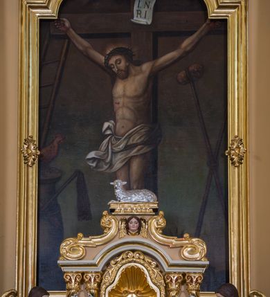 Zdjęcie nr 4: Jednokondygnacyjna nastawa ze zwieńczeniem uzyskała architektoniczną strukturę wzbogaconą o  flankujące ją bramkami. Współczesny marmurowy stipes ołtarzowy wspierający mensę uzyskał formę prostopadłościanu z licem artykułowanym dwiema półkolumienkami. Na mensie ustawiono tabernakulum w formie drewnianej skrzynki, obitej trybowaną blachą. Drzwiczki uzyskały kształt stojącego prostokąta zamkniętego łukiem półkolistym. W ich polu wyobrażono kielich z hostią w glorii, ujęty trybowanym winnym gronem i aplikowanymi kłosami zbóż. Z dwóch stron drzwiczek widnieje trybowana dekoracja w postaci winnego grona. Na tabernakulum usytuowano tron wystawienia, w postaci obrotowej niszy ujętej dwiema parami kolumienek podtrzymujących gzyms wyłamany pośrodku na kształt łuku odcinkowego oraz cokół z figurką baranka na księdze z siedmioma pieczęciami, podtrzymywany przez dwa spływy wolutowe. Tabernakulum ujęte zostało dwoma postumentami dekorowanymi z przodu rozetami. Ustawiono na nich figury klęczących aniołów. Nastawa ujęta została dwiema parami kolumn o kapitelach kompozytowych ustawionych na cokołach dekorowanych płycinami z ornamentem roślinnym, podtrzymujących pełne belkowanie z silnie wysuniętym gzymsem, oraz zewnętrzną parą cofniętych w tył kolumn. W polu głównym usytuowano obraz z przedstawieniem Zmartwychwstałego Chrystusa ze św. Janem Chrzcicielem i św. Janem Ewangelistą, ujęty profilowaną ramą w formie stojącego prostokąta zamkniętego łukiem odcinkowych nadwieszonym. Kondygnacja ujęta została po bokach uszami utworzonymi z karbowanej wstęgi i akantu. Zwieńczenie nastawy ustawiono na schodkowym, zwężającym się cokole, dekorowanym medalionem z monogramem Marii oraz anthemionami. Ujmują je dwie pary kolumienek o kapitelach kompozytowych podtrzymujących gzyms wyłamujący się po środku na kształt łuku półkolistego, dekorowany wicią roślinną. W polu zwieńczenia znalazł się obraz w formie stojącego owalu z przedstawieniem Dzieciątka Jezus prezentującego krzyż, ujęty profilowaną ramą dekorowaną ornamentem roślinnym. Zwieńczenie flankują stojące na gzymsie figury św. Józefa z Dzieciątkiem i Symeona z Dzieciątkiem. Nastawę flankują znajdujące się przy ścianie para kolumn i para półkolumn ustawionych na dwustrefowych cokołach, dźwigających pełne belkowanie, na którym siedzą dwa putta. Pomiędzy cokołem nastawy i zewnętrznych podpór ulokowano wygięte listwy tworzące bramki, na których stoją figury św. Piotra i św. Pawła. Struktura retabulum jest polichromowana w kolorze beżowym, z białymi i czerwonymi profilami i płycinami. Kolumny, ornamentyka i profile zostały pozłocone.