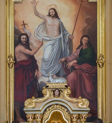 Zdjęcie nr 20: Jednokondygnacyjna nastawa ze zwieńczeniem uzyskała architektoniczną strukturę wzbogaconą o  flankujące ją bramkami. Współczesny marmurowy stipes ołtarzowy wspierający mensę uzyskał formę prostopadłościanu z licem artykułowanym dwiema półkolumienkami. Na mensie ustawiono tabernakulum w formie drewnianej skrzynki, obitej trybowaną blachą. Drzwiczki uzyskały kształt stojącego prostokąta zamkniętego łukiem półkolistym. W ich polu wyobrażono kielich z hostią w glorii, ujęty trybowanym winnym gronem i aplikowanymi kłosami zbóż. Z dwóch stron drzwiczek widnieje trybowana dekoracja w postaci winnego grona. Na tabernakulum usytuowano tron wystawienia, w postaci obrotowej niszy ujętej dwiema parami kolumienek podtrzymujących gzyms wyłamany pośrodku na kształt łuku odcinkowego oraz cokół z figurką baranka na księdze z siedmioma pieczęciami, podtrzymywany przez dwa spływy wolutowe. Tabernakulum ujęte zostało dwoma postumentami dekorowanymi z przodu rozetami. Ustawiono na nich figury klęczących aniołów. Nastawa ujęta została dwiema parami kolumn o kapitelach kompozytowych ustawionych na cokołach dekorowanych płycinami z ornamentem roślinnym, podtrzymujących pełne belkowanie z silnie wysuniętym gzymsem, oraz zewnętrzną parą cofniętych w tył kolumn. W polu głównym usytuowano obraz z przedstawieniem Zmartwychwstałego Chrystusa ze św. Janem Chrzcicielem i św. Janem Ewangelistą, ujęty profilowaną ramą w formie stojącego prostokąta zamkniętego łukiem odcinkowych nadwieszonym. Kondygnacja ujęta została po bokach uszami utworzonymi z karbowanej wstęgi i akantu. Zwieńczenie nastawy ustawiono na schodkowym, zwężającym się cokole, dekorowanym medalionem z monogramem Marii oraz anthemionami. Ujmują je dwie pary kolumienek o kapitelach kompozytowych podtrzymujących gzyms wyłamujący się po środku na kształt łuku półkolistego, dekorowany wicią roślinną. W polu zwieńczenia znalazł się obraz w formie stojącego owalu z przedstawieniem Dzieciątka Jezus prezentującego krzyż, ujęty profilowaną ramą dekorowaną ornamentem roślinnym. Zwieńczenie flankują stojące na gzymsie figury św. Józefa z Dzieciątkiem i Symeona z Dzieciątkiem. Nastawę flankują znajdujące się przy ścianie para kolumn i para półkolumn ustawionych na dwustrefowych cokołach, dźwigających pełne belkowanie, na którym siedzą dwa putta. Pomiędzy cokołem nastawy i zewnętrznych podpór ulokowano wygięte listwy tworzące bramki, na których stoją figury św. Piotra i św. Pawła. Struktura retabulum jest polichromowana w kolorze beżowym, z białymi i czerwonymi profilami i płycinami. Kolumny, ornamentyka i profile zostały pozłocone.