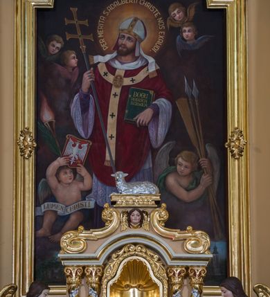 Zdjęcie nr 24: Jednokondygnacyjna nastawa ze zwieńczeniem uzyskała architektoniczną strukturę wzbogaconą o  flankujące ją bramkami. Współczesny marmurowy stipes ołtarzowy wspierający mensę uzyskał formę prostopadłościanu z licem artykułowanym dwiema półkolumienkami. Na mensie ustawiono tabernakulum w formie drewnianej skrzynki, obitej trybowaną blachą. Drzwiczki uzyskały kształt stojącego prostokąta zamkniętego łukiem półkolistym. W ich polu wyobrażono kielich z hostią w glorii, ujęty trybowanym winnym gronem i aplikowanymi kłosami zbóż. Z dwóch stron drzwiczek widnieje trybowana dekoracja w postaci winnego grona. Na tabernakulum usytuowano tron wystawienia, w postaci obrotowej niszy ujętej dwiema parami kolumienek podtrzymujących gzyms wyłamany pośrodku na kształt łuku odcinkowego oraz cokół z figurką baranka na księdze z siedmioma pieczęciami, podtrzymywany przez dwa spływy wolutowe. Tabernakulum ujęte zostało dwoma postumentami dekorowanymi z przodu rozetami. Ustawiono na nich figury klęczących aniołów. Nastawa ujęta została dwiema parami kolumn o kapitelach kompozytowych ustawionych na cokołach dekorowanych płycinami z ornamentem roślinnym, podtrzymujących pełne belkowanie z silnie wysuniętym gzymsem, oraz zewnętrzną parą cofniętych w tył kolumn. W polu głównym usytuowano obraz z przedstawieniem Zmartwychwstałego Chrystusa ze św. Janem Chrzcicielem i św. Janem Ewangelistą, ujęty profilowaną ramą w formie stojącego prostokąta zamkniętego łukiem odcinkowych nadwieszonym. Kondygnacja ujęta została po bokach uszami utworzonymi z karbowanej wstęgi i akantu. Zwieńczenie nastawy ustawiono na schodkowym, zwężającym się cokole, dekorowanym medalionem z monogramem Marii oraz anthemionami. Ujmują je dwie pary kolumienek o kapitelach kompozytowych podtrzymujących gzyms wyłamujący się po środku na kształt łuku półkolistego, dekorowany wicią roślinną. W polu zwieńczenia znalazł się obraz w formie stojącego owalu z przedstawieniem Dzieciątka Jezus prezentującego krzyż, ujęty profilowaną ramą dekorowaną ornamentem roślinnym. Zwieńczenie flankują stojące na gzymsie figury św. Józefa z Dzieciątkiem i Symeona z Dzieciątkiem. Nastawę flankują znajdujące się przy ścianie para kolumn i para półkolumn ustawionych na dwustrefowych cokołach, dźwigających pełne belkowanie, na którym siedzą dwa putta. Pomiędzy cokołem nastawy i zewnętrznych podpór ulokowano wygięte listwy tworzące bramki, na których stoją figury św. Piotra i św. Pawła. Struktura retabulum jest polichromowana w kolorze beżowym, z białymi i czerwonymi profilami i płycinami. Kolumny, ornamentyka i profile zostały pozłocone.