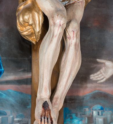 Zdjęcie nr 6: Malarsko-rzeźbiarska scena Ukrzyżowania w polu w kształcie stojącego prostokąta. Pełnoplastyczna figura Ukrzyżowanego w typie Christo morto. Głowa mocno pochylona w prawo, pociągła, wyraziście modelowana twarz o długim, prostym nosie, rozchylonych ustach i krótkim, ciemnobrązowym zaroście. Długie, falowane włosy opadają na prawe ramię. Głowę wieńczy wydatna, złocona korona. Ciało o wyrazistej, plastycznie opracowanej muskulaturze i delikatnie zaznaczonej linii żeber, z drobną fałdką pośrodku brzucha. Biodra przewiązane krótkim, silnie drapowanym, złoconym perizonium z długim zwisem na prawym boku. Zgięte w kolanach, lekko przechylone w lewo nogi przybite są 1 gwoździem. Na czole i klatce piersiowej drobnie ślady krwi, na rękach, prawym boku i stopach wydatne strugi. W górnej partii krzyża biały titulus w formie zwoju z napisem „I.N. / R.I”. Figury asystencyjne namalowane na tle rozległego pejzażu miasta i ciemnego nieba z zaćmionym słońce w lewym, górnym rogu. Po prawej stronie krzyża stoi Matka Boska skierowana w trzech czwartych w prawo odwracając się w za siebie. Owalna twarz o delikatnych rysach, oczy półprzymknięte. Ubrana w jasnoróżową suknię, narzucony na głowę ciemnoniebieski płaszcz i okalającą szyję białą chustę. Ręce złocone w geście modlitwy. Po lewej stronie krzyża stoi św. Jan Ewangelista zwrócony w trzech czwartych w prawo. Owalna twarz o delikatnych rysach, wzrok skierowany na wprost, z oczu płyną krople łez. Falowane włosy opadają na ramiona. Ubrany jest w zieloną, przewiązana w pasie suknię i bordowy, narzucony na ramiona płaszcz. na stopach sandały. Prawa ręka uniesiona na wysokości talii, lewa odsunięta w bok. Głowę Marii i sw. Jana okalają złocone nimby.   