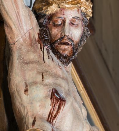 Zdjęcie nr 5: Malarsko-rzeźbiarska scena Ukrzyżowania w polu w kształcie stojącego prostokąta. Pełnoplastyczna figura Ukrzyżowanego w typie Christo morto. Głowa mocno pochylona w prawo, pociągła, wyraziście modelowana twarz o długim, prostym nosie, rozchylonych ustach i krótkim, ciemnobrązowym zaroście. Długie, falowane włosy opadają na prawe ramię. Głowę wieńczy wydatna, złocona korona. Ciało o wyrazistej, plastycznie opracowanej muskulaturze i delikatnie zaznaczonej linii żeber, z drobną fałdką pośrodku brzucha. Biodra przewiązane krótkim, silnie drapowanym, złoconym perizonium z długim zwisem na prawym boku. Zgięte w kolanach, lekko przechylone w lewo nogi przybite są 1 gwoździem. Na czole i klatce piersiowej drobnie ślady krwi, na rękach, prawym boku i stopach wydatne strugi. W górnej partii krzyża biały titulus w formie zwoju z napisem „I.N. / R.I”. Figury asystencyjne namalowane na tle rozległego pejzażu miasta i ciemnego nieba z zaćmionym słońce w lewym, górnym rogu. Po prawej stronie krzyża stoi Matka Boska skierowana w trzech czwartych w prawo odwracając się w za siebie. Owalna twarz o delikatnych rysach, oczy półprzymknięte. Ubrana w jasnoróżową suknię, narzucony na głowę ciemnoniebieski płaszcz i okalającą szyję białą chustę. Ręce złocone w geście modlitwy. Po lewej stronie krzyża stoi św. Jan Ewangelista zwrócony w trzech czwartych w prawo. Owalna twarz o delikatnych rysach, wzrok skierowany na wprost, z oczu płyną krople łez. Falowane włosy opadają na ramiona. Ubrany jest w zieloną, przewiązana w pasie suknię i bordowy, narzucony na ramiona płaszcz. na stopach sandały. Prawa ręka uniesiona na wysokości talii, lewa odsunięta w bok. Głowę Marii i sw. Jana okalają złocone nimby.   