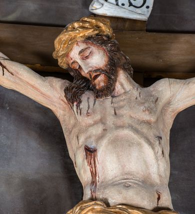 Zdjęcie nr 3: Malarsko-rzeźbiarska scena Ukrzyżowania w polu w kształcie stojącego prostokąta. Pełnoplastyczna figura Ukrzyżowanego w typie Christo morto. Głowa mocno pochylona w prawo, pociągła, wyraziście modelowana twarz o długim, prostym nosie, rozchylonych ustach i krótkim, ciemnobrązowym zaroście. Długie, falowane włosy opadają na prawe ramię. Głowę wieńczy wydatna, złocona korona. Ciało o wyrazistej, plastycznie opracowanej muskulaturze i delikatnie zaznaczonej linii żeber, z drobną fałdką pośrodku brzucha. Biodra przewiązane krótkim, silnie drapowanym, złoconym perizonium z długim zwisem na prawym boku. Zgięte w kolanach, lekko przechylone w lewo nogi przybite są 1 gwoździem. Na czole i klatce piersiowej drobnie ślady krwi, na rękach, prawym boku i stopach wydatne strugi. W górnej partii krzyża biały titulus w formie zwoju z napisem „I.N. / R.I”. Figury asystencyjne namalowane na tle rozległego pejzażu miasta i ciemnego nieba z zaćmionym słońce w lewym, górnym rogu. Po prawej stronie krzyża stoi Matka Boska skierowana w trzech czwartych w prawo odwracając się w za siebie. Owalna twarz o delikatnych rysach, oczy półprzymknięte. Ubrana w jasnoróżową suknię, narzucony na głowę ciemnoniebieski płaszcz i okalającą szyję białą chustę. Ręce złocone w geście modlitwy. Po lewej stronie krzyża stoi św. Jan Ewangelista zwrócony w trzech czwartych w prawo. Owalna twarz o delikatnych rysach, wzrok skierowany na wprost, z oczu płyną krople łez. Falowane włosy opadają na ramiona. Ubrany jest w zieloną, przewiązana w pasie suknię i bordowy, narzucony na ramiona płaszcz. na stopach sandały. Prawa ręka uniesiona na wysokości talii, lewa odsunięta w bok. Głowę Marii i sw. Jana okalają złocone nimby.   