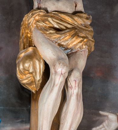 Zdjęcie nr 7: Malarsko-rzeźbiarska scena Ukrzyżowania w polu w kształcie stojącego prostokąta. Pełnoplastyczna figura Ukrzyżowanego w typie Christo morto. Głowa mocno pochylona w prawo, pociągła, wyraziście modelowana twarz o długim, prostym nosie, rozchylonych ustach i krótkim, ciemnobrązowym zaroście. Długie, falowane włosy opadają na prawe ramię. Głowę wieńczy wydatna, złocona korona. Ciało o wyrazistej, plastycznie opracowanej muskulaturze i delikatnie zaznaczonej linii żeber, z drobną fałdką pośrodku brzucha. Biodra przewiązane krótkim, silnie drapowanym, złoconym perizonium z długim zwisem na prawym boku. Zgięte w kolanach, lekko przechylone w lewo nogi przybite są 1 gwoździem. Na czole i klatce piersiowej drobnie ślady krwi, na rękach, prawym boku i stopach wydatne strugi. W górnej partii krzyża biały titulus w formie zwoju z napisem „I.N. / R.I”. Figury asystencyjne namalowane na tle rozległego pejzażu miasta i ciemnego nieba z zaćmionym słońce w lewym, górnym rogu. Po prawej stronie krzyża stoi Matka Boska skierowana w trzech czwartych w prawo odwracając się w za siebie. Owalna twarz o delikatnych rysach, oczy półprzymknięte. Ubrana w jasnoróżową suknię, narzucony na głowę ciemnoniebieski płaszcz i okalającą szyję białą chustę. Ręce złocone w geście modlitwy. Po lewej stronie krzyża stoi św. Jan Ewangelista zwrócony w trzech czwartych w prawo. Owalna twarz o delikatnych rysach, wzrok skierowany na wprost, z oczu płyną krople łez. Falowane włosy opadają na ramiona. Ubrany jest w zieloną, przewiązana w pasie suknię i bordowy, narzucony na ramiona płaszcz. na stopach sandały. Prawa ręka uniesiona na wysokości talii, lewa odsunięta w bok. Głowę Marii i sw. Jana okalają złocone nimby.   