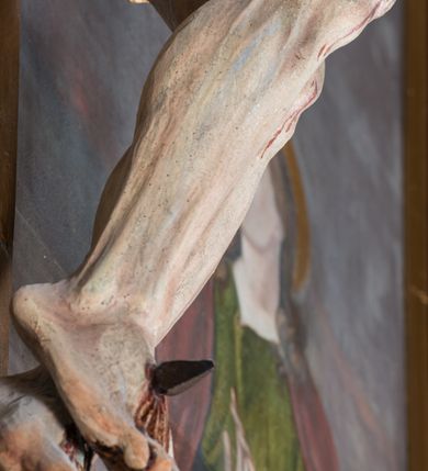 Zdjęcie nr 10: Malarsko-rzeźbiarska scena Ukrzyżowania w polu w kształcie stojącego prostokąta. Pełnoplastyczna figura Ukrzyżowanego w typie Christo morto. Głowa mocno pochylona w prawo, pociągła, wyraziście modelowana twarz o długim, prostym nosie, rozchylonych ustach i krótkim, ciemnobrązowym zaroście. Długie, falowane włosy opadają na prawe ramię. Głowę wieńczy wydatna, złocona korona. Ciało o wyrazistej, plastycznie opracowanej muskulaturze i delikatnie zaznaczonej linii żeber, z drobną fałdką pośrodku brzucha. Biodra przewiązane krótkim, silnie drapowanym, złoconym perizonium z długim zwisem na prawym boku. Zgięte w kolanach, lekko przechylone w lewo nogi przybite są 1 gwoździem. Na czole i klatce piersiowej drobnie ślady krwi, na rękach, prawym boku i stopach wydatne strugi. W górnej partii krzyża biały titulus w formie zwoju z napisem „I.N. / R.I”. Figury asystencyjne namalowane na tle rozległego pejzażu miasta i ciemnego nieba z zaćmionym słońce w lewym, górnym rogu. Po prawej stronie krzyża stoi Matka Boska skierowana w trzech czwartych w prawo odwracając się w za siebie. Owalna twarz o delikatnych rysach, oczy półprzymknięte. Ubrana w jasnoróżową suknię, narzucony na głowę ciemnoniebieski płaszcz i okalającą szyję białą chustę. Ręce złocone w geście modlitwy. Po lewej stronie krzyża stoi św. Jan Ewangelista zwrócony w trzech czwartych w prawo. Owalna twarz o delikatnych rysach, wzrok skierowany na wprost, z oczu płyną krople łez. Falowane włosy opadają na ramiona. Ubrany jest w zieloną, przewiązana w pasie suknię i bordowy, narzucony na ramiona płaszcz. na stopach sandały. Prawa ręka uniesiona na wysokości talii, lewa odsunięta w bok. Głowę Marii i sw. Jana okalają złocone nimby.   