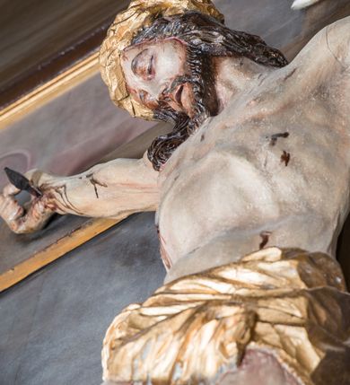 Zdjęcie nr 4: Malarsko-rzeźbiarska scena Ukrzyżowania w polu w kształcie stojącego prostokąta. Pełnoplastyczna figura Ukrzyżowanego w typie Christo morto. Głowa mocno pochylona w prawo, pociągła, wyraziście modelowana twarz o długim, prostym nosie, rozchylonych ustach i krótkim, ciemnobrązowym zaroście. Długie, falowane włosy opadają na prawe ramię. Głowę wieńczy wydatna, złocona korona. Ciało o wyrazistej, plastycznie opracowanej muskulaturze i delikatnie zaznaczonej linii żeber, z drobną fałdką pośrodku brzucha. Biodra przewiązane krótkim, silnie drapowanym, złoconym perizonium z długim zwisem na prawym boku. Zgięte w kolanach, lekko przechylone w lewo nogi przybite są 1 gwoździem. Na czole i klatce piersiowej drobnie ślady krwi, na rękach, prawym boku i stopach wydatne strugi. W górnej partii krzyża biały titulus w formie zwoju z napisem „I.N. / R.I”. Figury asystencyjne namalowane na tle rozległego pejzażu miasta i ciemnego nieba z zaćmionym słońce w lewym, górnym rogu. Po prawej stronie krzyża stoi Matka Boska skierowana w trzech czwartych w prawo odwracając się w za siebie. Owalna twarz o delikatnych rysach, oczy półprzymknięte. Ubrana w jasnoróżową suknię, narzucony na głowę ciemnoniebieski płaszcz i okalającą szyję białą chustę. Ręce złocone w geście modlitwy. Po lewej stronie krzyża stoi św. Jan Ewangelista zwrócony w trzech czwartych w prawo. Owalna twarz o delikatnych rysach, wzrok skierowany na wprost, z oczu płyną krople łez. Falowane włosy opadają na ramiona. Ubrany jest w zieloną, przewiązana w pasie suknię i bordowy, narzucony na ramiona płaszcz. na stopach sandały. Prawa ręka uniesiona na wysokości talii, lewa odsunięta w bok. Głowę Marii i sw. Jana okalają złocone nimby.   