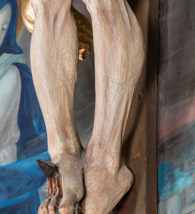 Zdjęcie nr 9: Malarsko-rzeźbiarska scena Ukrzyżowania w polu w kształcie stojącego prostokąta. Pełnoplastyczna figura Ukrzyżowanego w typie Christo morto. Głowa mocno pochylona w prawo, pociągła, wyraziście modelowana twarz o długim, prostym nosie, rozchylonych ustach i krótkim, ciemnobrązowym zaroście. Długie, falowane włosy opadają na prawe ramię. Głowę wieńczy wydatna, złocona korona. Ciało o wyrazistej, plastycznie opracowanej muskulaturze i delikatnie zaznaczonej linii żeber, z drobną fałdką pośrodku brzucha. Biodra przewiązane krótkim, silnie drapowanym, złoconym perizonium z długim zwisem na prawym boku. Zgięte w kolanach, lekko przechylone w lewo nogi przybite są 1 gwoździem. Na czole i klatce piersiowej drobnie ślady krwi, na rękach, prawym boku i stopach wydatne strugi. W górnej partii krzyża biały titulus w formie zwoju z napisem „I.N. / R.I”. Figury asystencyjne namalowane na tle rozległego pejzażu miasta i ciemnego nieba z zaćmionym słońce w lewym, górnym rogu. Po prawej stronie krzyża stoi Matka Boska skierowana w trzech czwartych w prawo odwracając się w za siebie. Owalna twarz o delikatnych rysach, oczy półprzymknięte. Ubrana w jasnoróżową suknię, narzucony na głowę ciemnoniebieski płaszcz i okalającą szyję białą chustę. Ręce złocone w geście modlitwy. Po lewej stronie krzyża stoi św. Jan Ewangelista zwrócony w trzech czwartych w prawo. Owalna twarz o delikatnych rysach, wzrok skierowany na wprost, z oczu płyną krople łez. Falowane włosy opadają na ramiona. Ubrany jest w zieloną, przewiązana w pasie suknię i bordowy, narzucony na ramiona płaszcz. na stopach sandały. Prawa ręka uniesiona na wysokości talii, lewa odsunięta w bok. Głowę Marii i sw. Jana okalają złocone nimby.   