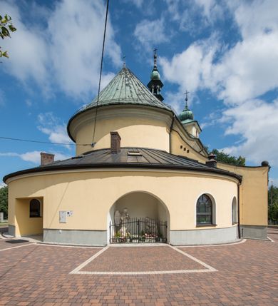 Zdjęcie nr 16: Orientowany kościół św. Wojciecha w Bulowicach obecnie uzyskał strukturę trójnawowej, trójprzęsłowej bazyliki z jednoprzęsłowym, nieco węższym prezbiterium zamkniętym spłaszczoną absydą. Na przedłużeniu dostawionych naw wybudowano półkolisty aneks zakrystyjny okalający apsydę, przepruwając go trzema półokrągłymi oknami oraz wejściem na osi. W aneksie od strony południowo-wschodniej urządzono otwartą na zewnątrz półkolistą arkadą kaplicę Ogrójcową. Od zachodu do nawy głównej przylega lekko wysunięta do przodu czworoboczna wieża flankowana dwoma, piętrowymi aneksami. Elewację masywu wieżowego artykułuje profilowany gzyms koronujący wyłamany na szerokości wieży, podtrzymujący miedziany daszek pulpitowy, dzielący ją na dwie kondygnacje. Podobny gzyms zamyka wieżę u góry, ponad którym wznosi się cebulasty hełm zwieńczony krzyżem na bani. Pod gzymsem w górnej części wieży usytuowano tarcze zegarowe oraz poniżej półokrągłe okna. W jej przyziemiu urządzono kruchtę, do której prowadzi portal uszaty z widocznym nad nim odcinkiem belkowania. Za pomocą symetrycznych spływów dostawione dwie nawy zostały połączone w spójną całość z masywem wieżowym. Do obu naw prowadzą półokrągłe arkady, w których urządzono otwarte przedsionki poprzedzające weście do kościoła. Nawę główną i prezbiterium nakryto dachem siodłowym. W jej części wschodniej nadwieszono wieżyczkę na sygnaturkę w kształcie sześciobocznej latarnii z baniastym hełmem i krzyżem. Nawy boczne i aneks zakrystyjny przykryto dachami pulpitowymi. Elewacje zewnętrzne zostały pozbawione artykulacji za wyjątkiem niskiego kamiennego cokołu obiegającego całą świątynię. Wnętrze nawy głównej zostało rozczłonkowane za pomocą opilastrowanych filarów, ponad którymi widoczne są odcinki belkowania. W tęczy kościoła zastosowano pilastry zdwojone. Nawy boczne połączono z nawą główną za pomocą mocno spłaszczonych arkad. Ponad nimi przepruto prostokątne okna zamknięte łukiem odcinkowym. Całość nawy nakryto sklepieniem żagielkowym wspartym na gurtach, zaś prezbiterium przykryto hemisferycznie. W części zachodniej nawy głównej zawieszono chór muzyczny wsparty na dwóch drewnianych filarach z drewnianym, pełnym parapetem artykułowanym czworobocznymi płycinami. Na osi środkowej parapet wyłamano na rzucie trapezu. W prezbiterium ustawiono ołtarz główny, a przy łuku tęczowym dwa ołtarze boczne. Na północnej ścianie łuku tęczowego zawieszono ambonę. Dwa niewielkie ołtarze boczne znalazły się także na zakończeniu naw bocznych.   