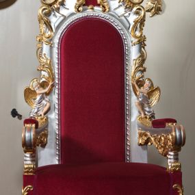 Zdjęcie nr 1: Fotel z wyściełanym wysokim oparciem i siedziskiem oraz dwiema poręczami z wyściełanymi podłokietnikami. Nóżki esowato wygięte, dekorowane ornamentem rocaille'owym obiegającym siedzisko u dołu. Tarcza oparcia w zwieńczeniu również zdobiona ornamentem rocaille'owym, spływającym aż na podłokietniki. Zaplecek flankowany parą aniołów, usadzonych na podłokietnikach z rękami wzniesionymi ku górze. Stelaż srebrzony ze złoconymi ornamentami, szaty aniołów srebrzone, skrzydła złocone, polichromia w odsłoniętych partiach ciała naturalistyczna. Tapicerka wtórna z bordowego aksamitu.




