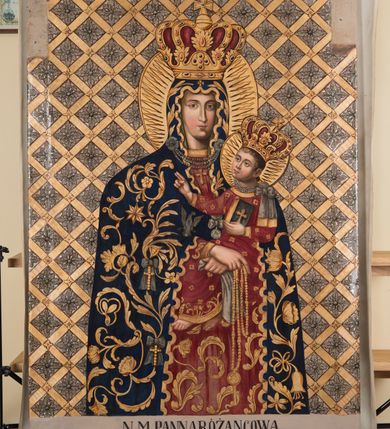 Zdjęcie nr 2: Obraz Matki Boskiej Różańcowej uzyskał kształt wydłużonego, stojącego prostokąta. Wizerunek przedstawia w półpostaci Matkę Boską z Dzieciątkiem. Maria, zwrócona delikatnie w trzech czwartych w lewo, podtrzymuje małego Jezusa obiema, skrzyżowanymi rękami na lewym ramieniu. Równocześnie Madonna w prawej dłoni trzyma białą chustę, a w lewej złoty różaniec zakończony owalnym medalionem z monogramem maryjnym. Jej rumiana twarz o jasnej karnacji jest modelowana miękkim światłocieniem. Twarz jest pociągła o delikatnych, łagodnych rysach, z prostym nosem i pełnych ustach. Matka Boska ubrana jest w ciemnogranatowy maforion podbity zielenią, lamowany i dekorowany złotymi motywami roślinno-kwiatowymi oraz imitacją zawieszonych wotów - białego orła, złotych krzyży z kokardami oraz srebrnego medalionu z kokardą. Maforion opadając na ramiona odsłania spodnią, szkarłatną suknię. Maria ma na głowie wysoką, złotą, zamkniętą koronę wypełnioną purpurą, zwieńczoną globem z krzyżykiem. Korona jest dekorowana malowaną kameryzacją. Pod szyją namalowano trzy sznury pereł przypiętych do maforiona dwiema spinkami w kształcie czteropłatkowych kwiatów. Na dolnym sznurze wisi złoty krzyżyk. Trzymane przez Madonnę Dzieciątko jest zwrócone w trzech czwartych w prawo. Mały Jezus lewą ręka podtrzymuje przy boku księgę Ewangelii z czarną okładką zdobioną złotym krzyżykiem. Natomiast nieznacznie usztywnioną i wyciągniętą przed siebie prawą rękę unosi w geście błogosławieństwa. Jezus ma okrągłą twarz o jasnej karnacji, pełnych ustach i rumianych policzkach, okoloną jasnobrązowymi, krótkimi włosami. Ubrany jest w szkarłatną tunikę u dołu bogato lamowaną złotym ornamentem roślinnym. Tunika pokryta jest drobnymi, złotymi, czterolistnymi kwiatuszkami. Na głowie ma wysoką, zamknietą, złotą koronę zwieńczoną krzyżykiem, dekorowaną malowaną kameryzacją i wypełnioną purpurą. Przy szyi małego Jezusa namalowano trzy krótkie sznury pereł dekorowane z lewej strony srebrną kokardą z długimi wstęgami. Głowy obu postaci otaczają złote nimby z wpisanymi w nie promieniami. Postacie zostały ukazane na srebrnym tle pokrytym złotą, romboidalną kratownicą. Pola kratownicy wypełniają rozety, a w miejscu przecięcia listew namalowano wielolistne kwiaty.
W dolnej części obrazu na białym tle umieszczono czarny napis: N(AJŚWIĘTSZA) M(ARIA) PANNA RÓŻAŃCOWA / Według obrazu cudami słynącego w kościele XX. Dominikanów w Krakowie / poświęconego przez Klemensa VIII Papieża a przywiezionego przez Kard(dynała) Ber / narda Maciejowskiego Bisk(upa) Krak(owskiego) Dobrodziejom spalonego kościoła S(więtej) Trójcy / R(oku) P(ańskiego) 1850. Pamiątka R(oku) P(ańskiego) 1862 malowany R(oku) P(ańskiego) 1866.